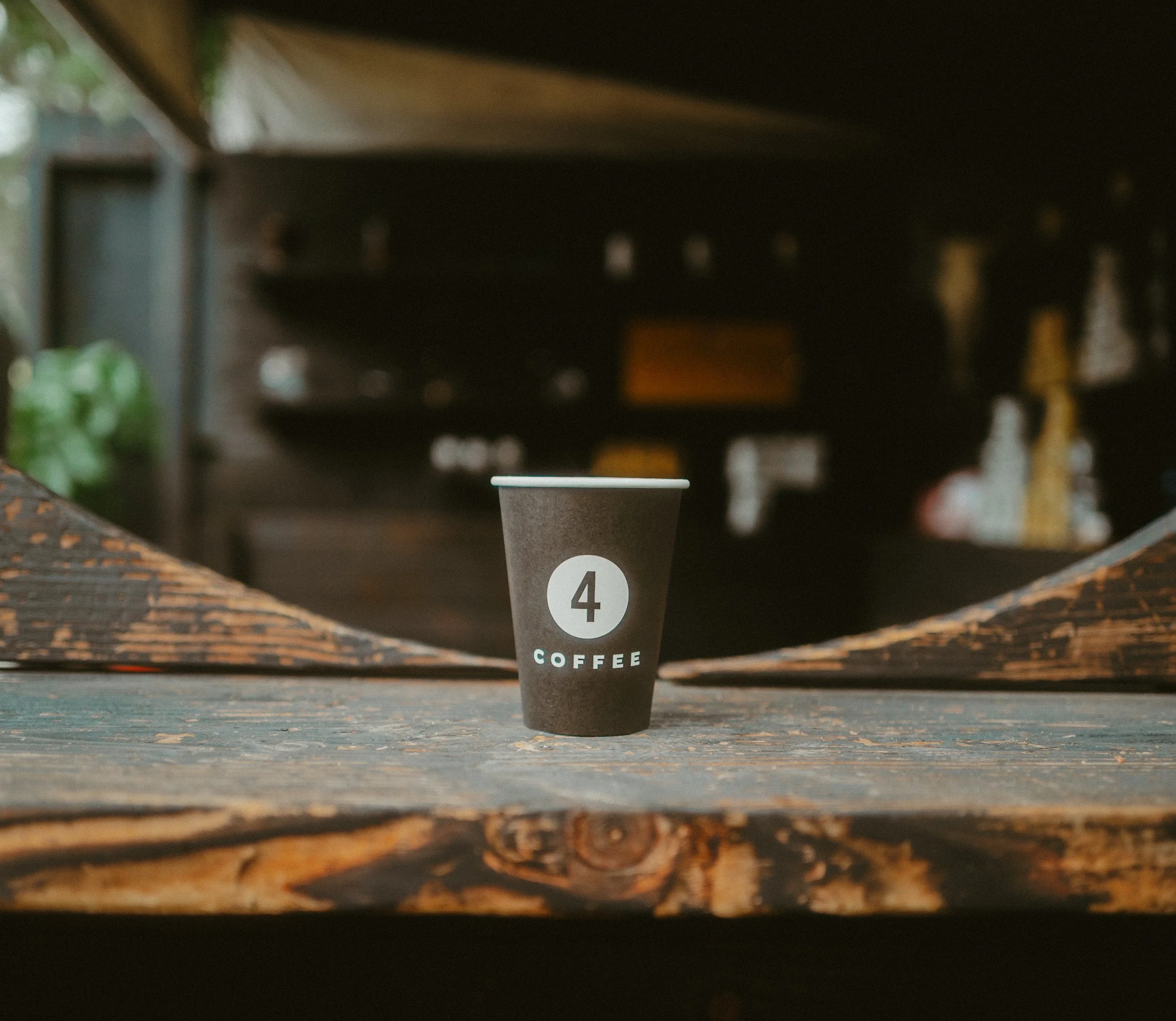 A black coffee cup with a white circle containing the number 4 and the word 'COFFEE' underneath, placed on a rustic wooden surface with a blurred store or cafe background.