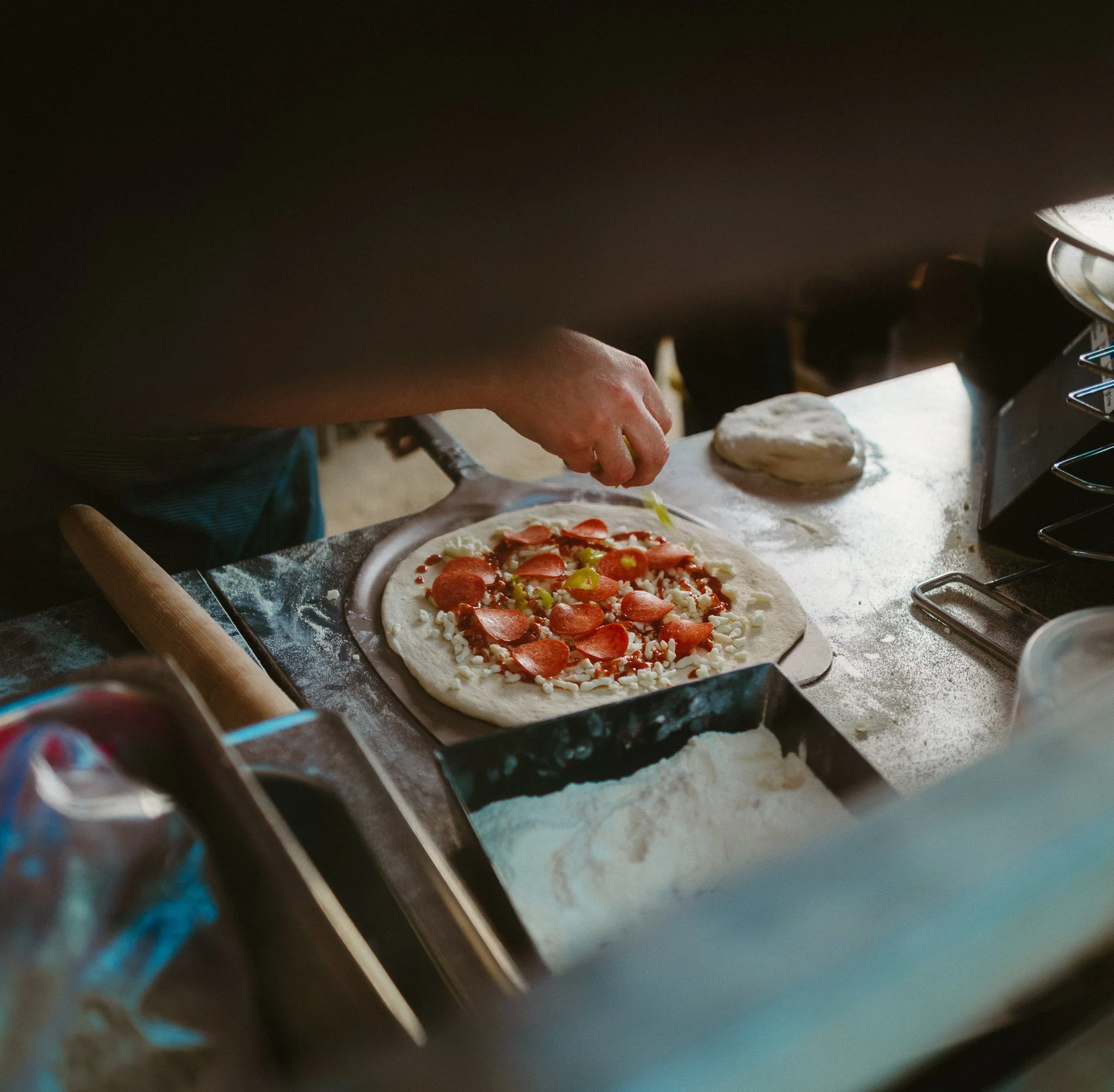 Person making a pizza, adding toppings like pepperoni and cheese on an uncooked pizza crust in a kitchen.
