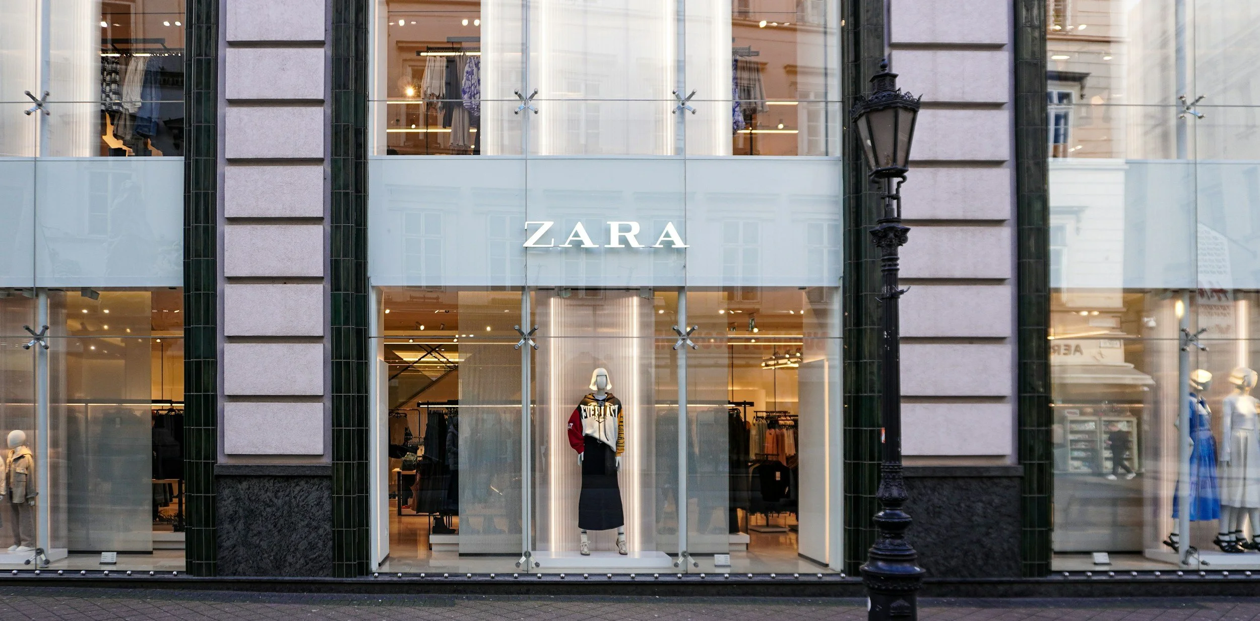 Front view of a Zara clothing store with mannequins displaying outfits, glass storefront showcasing clothing inside, and a vintage street lamp outside on the sidewalk.