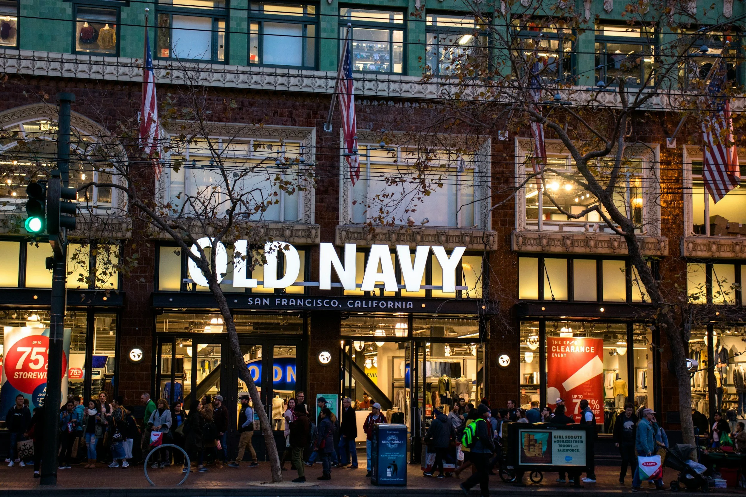 Exterior of a Old Navy store in San Francisco, California, with people walking on the sidewalk and American flags displayed outside.