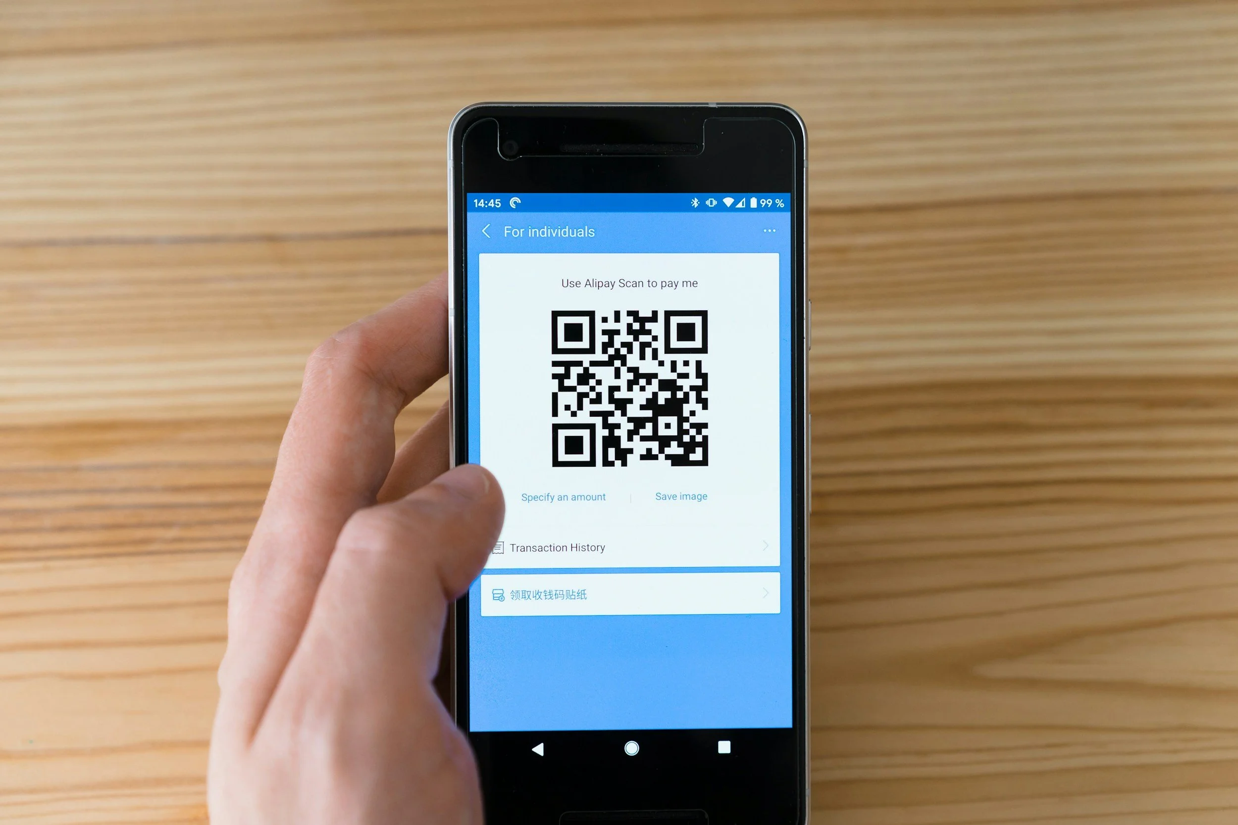A person holding a smartphone displaying a QR code on the screen, with a wooden surface in the background.