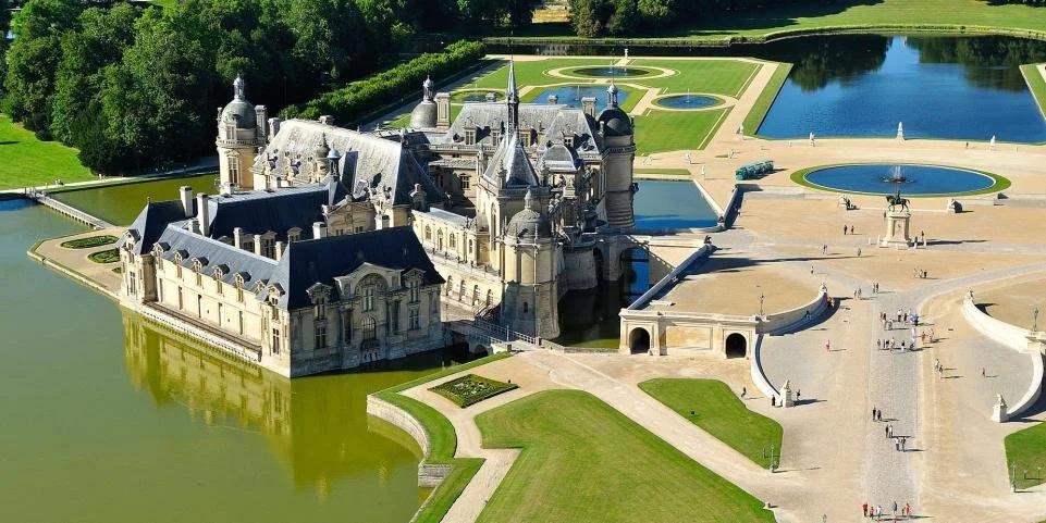 Aerial view of a large castle surrounded by water, with formal gardens and water features in the background.