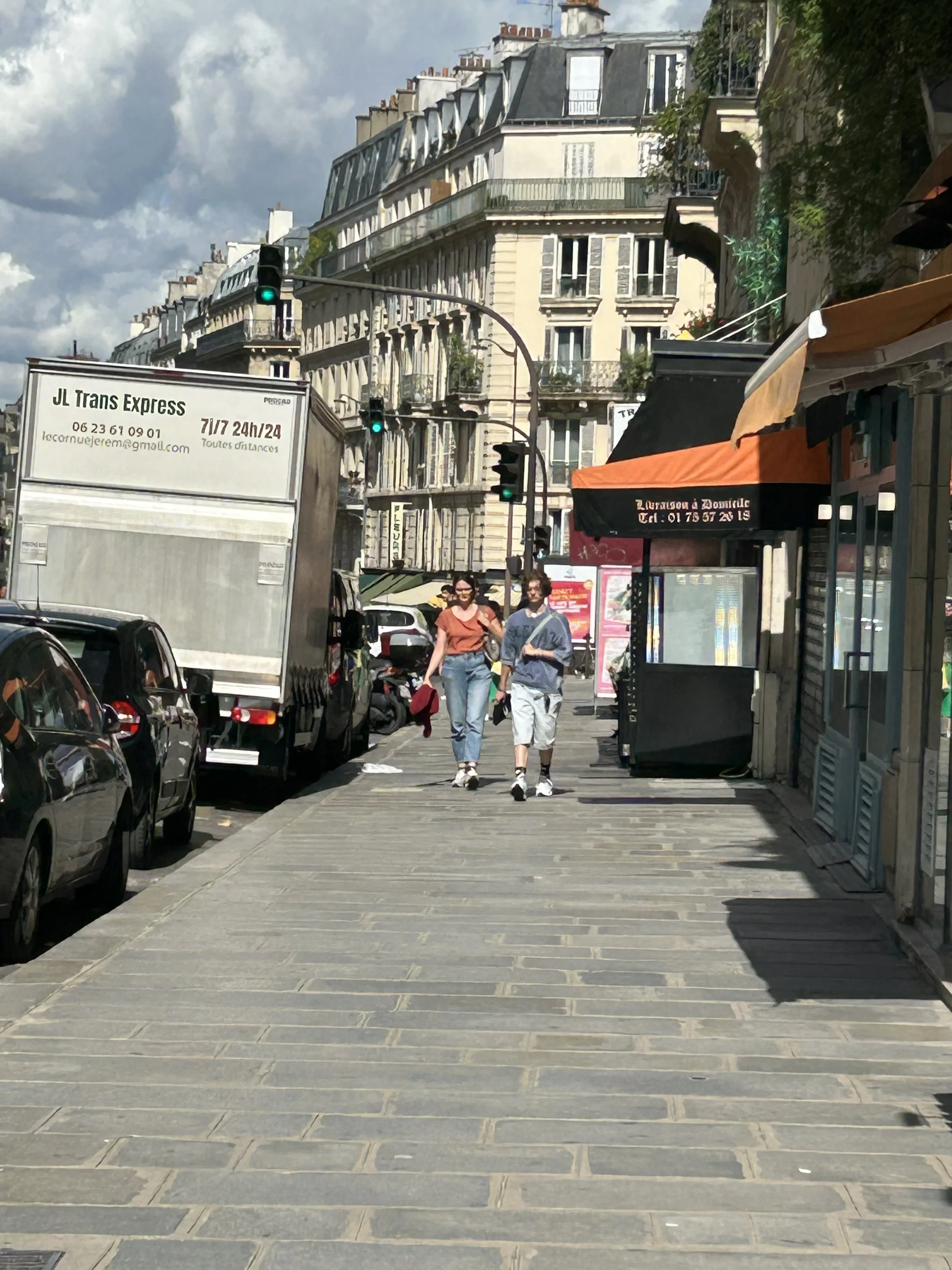 Paris street scene with pedestrians walking on a wide sidewalk, parked cars, building with classic Parisian architecture, and a delivery truck.