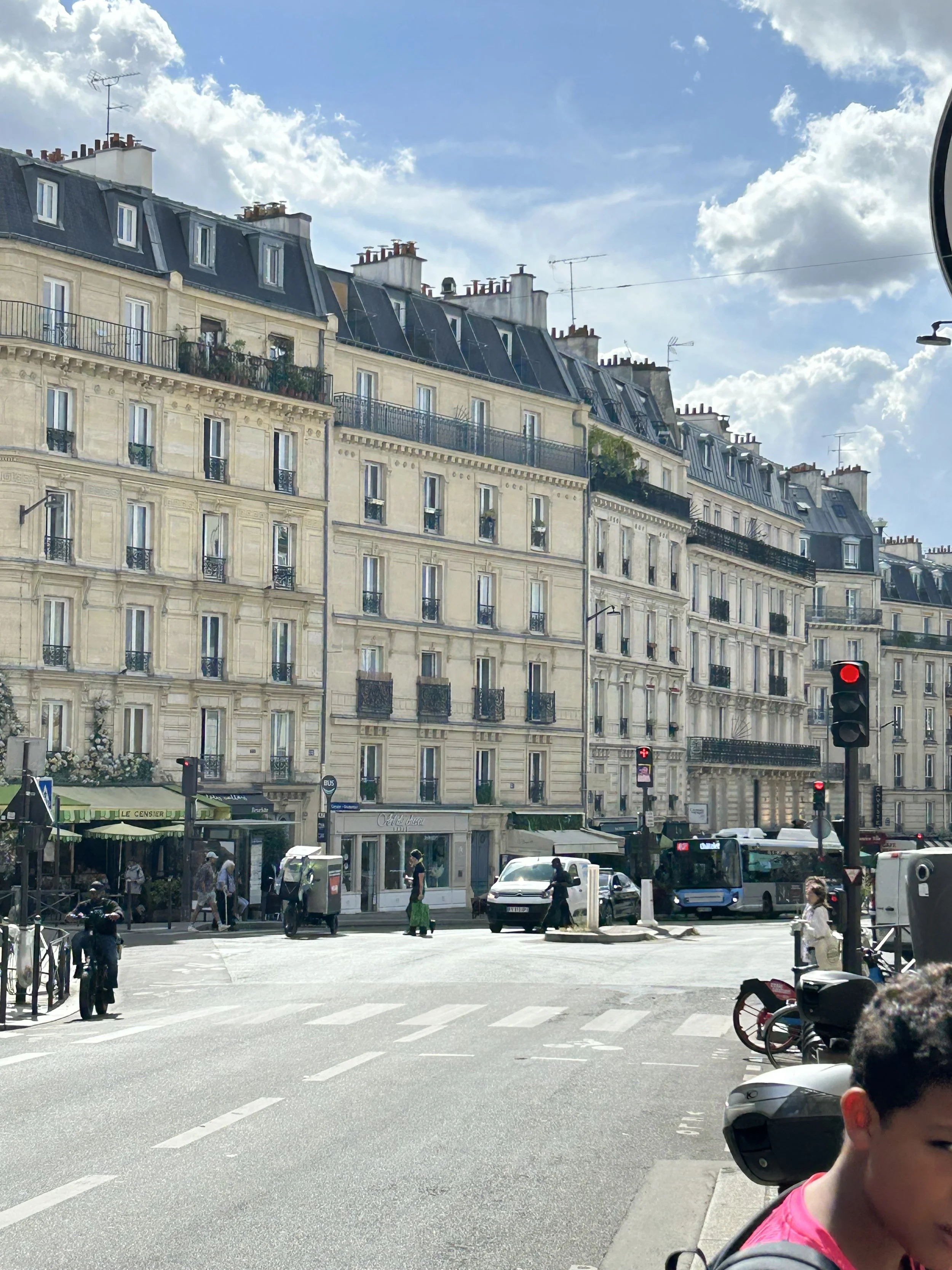 Parisian street scene with multi-story beige buildings featuring balconies, cars, a bus, pedestrians, and a traffic light