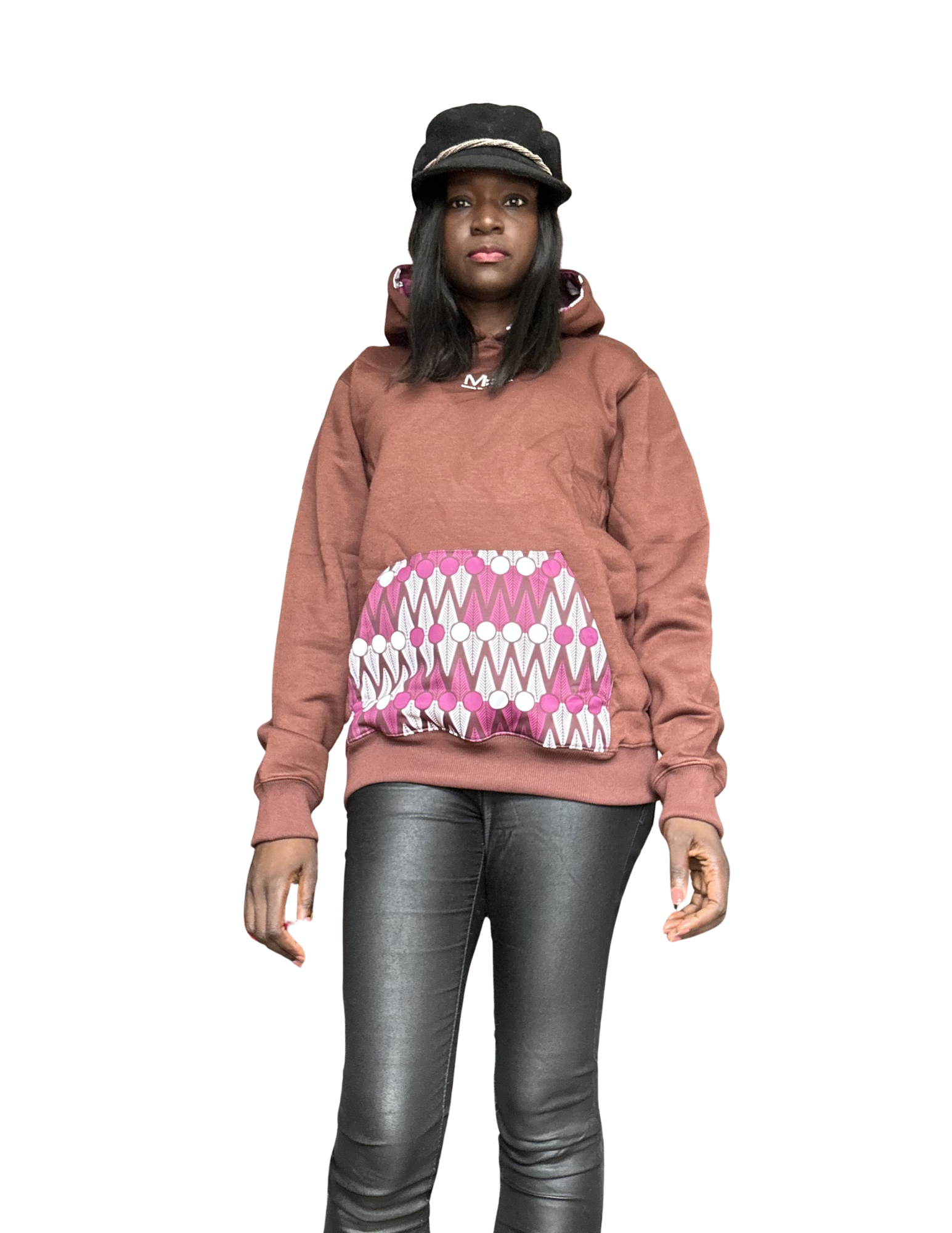 A woman standing against a plain white background, wearing a brown hoodie, black cap, and black leather pants.