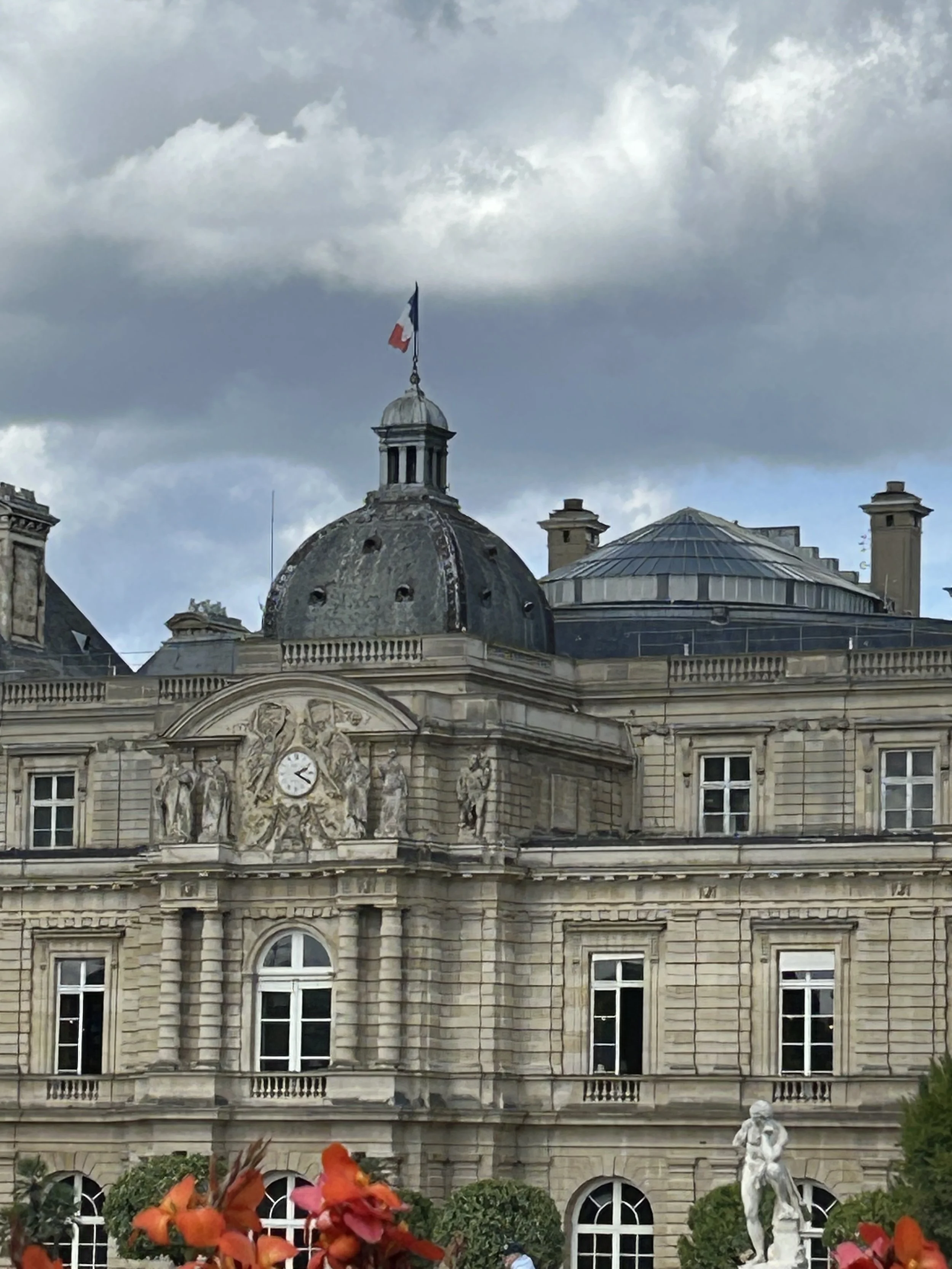 Historical building with ornate stone architecture, a clock, statues, and a French flag on top, under a cloudy sky.