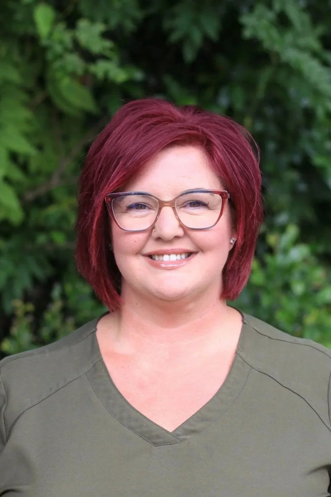A woman with short, vibrant red hair, wearing glasses with a light-colored frame, smiling, and standing outdoors in front of green foliage, wearing an olive green top.