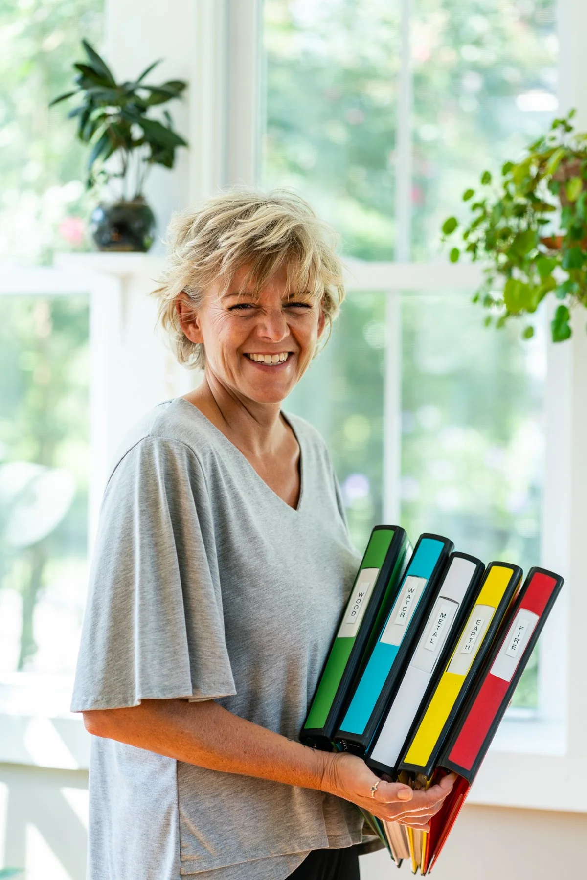 Smiling woman with short blonde hair holding five labeled binders in an indoor setting with a large window and plants in the background.