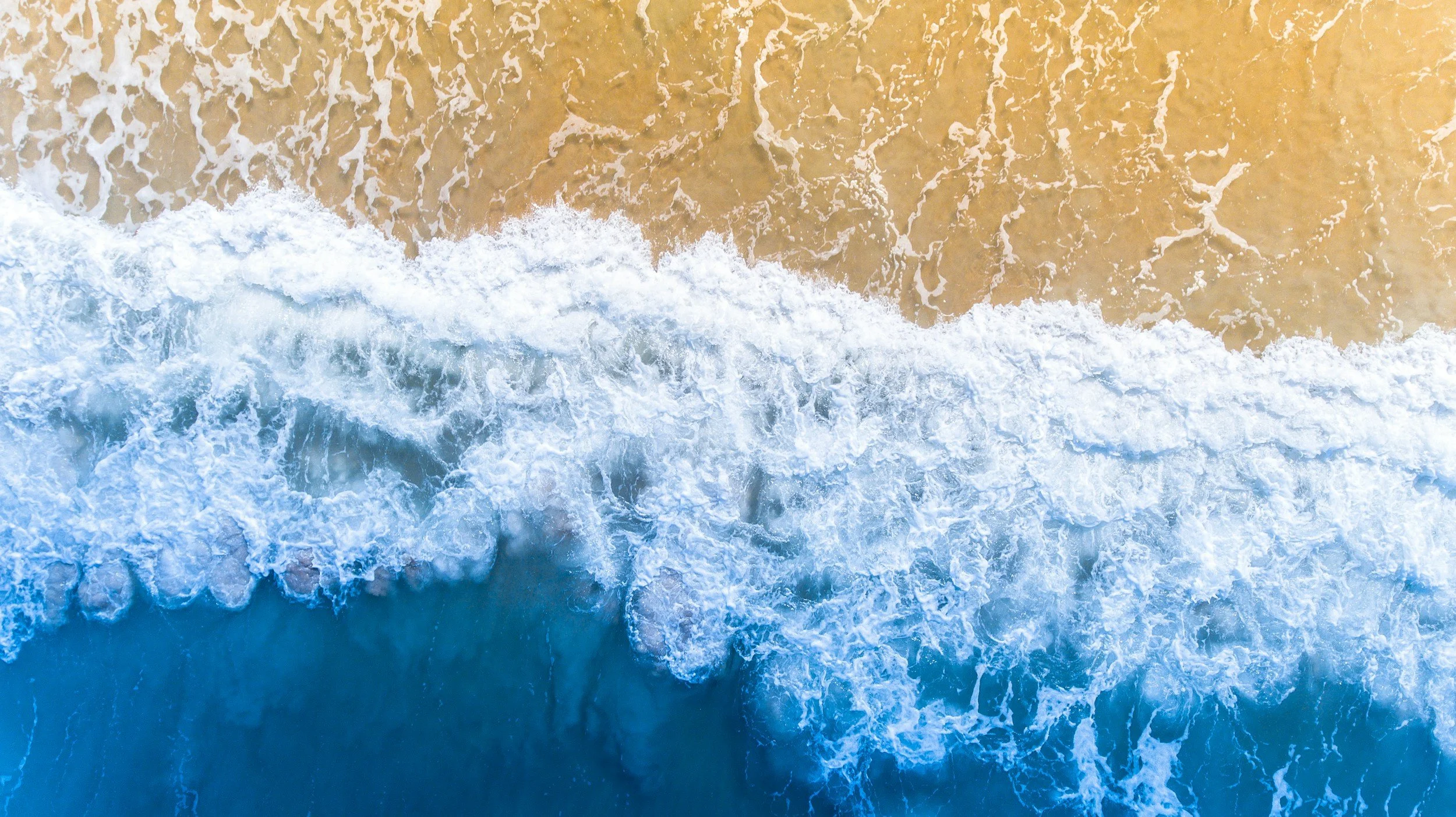 Overhead view of ocean waves crashing onto a sandy beach during daytime.
