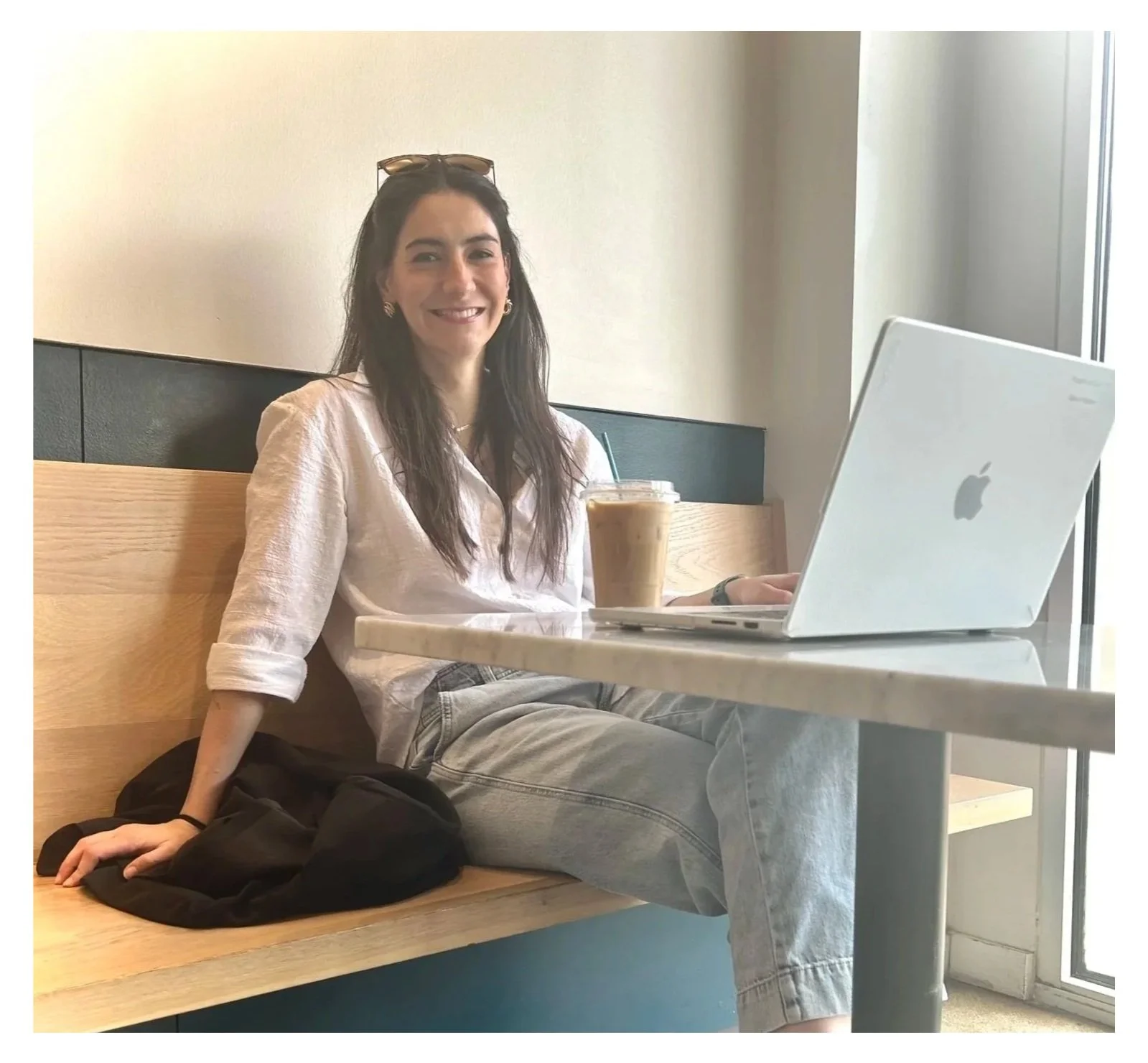 A woman sitting in a cafe with a silver laptop and iced coffee, smiling at the camera.