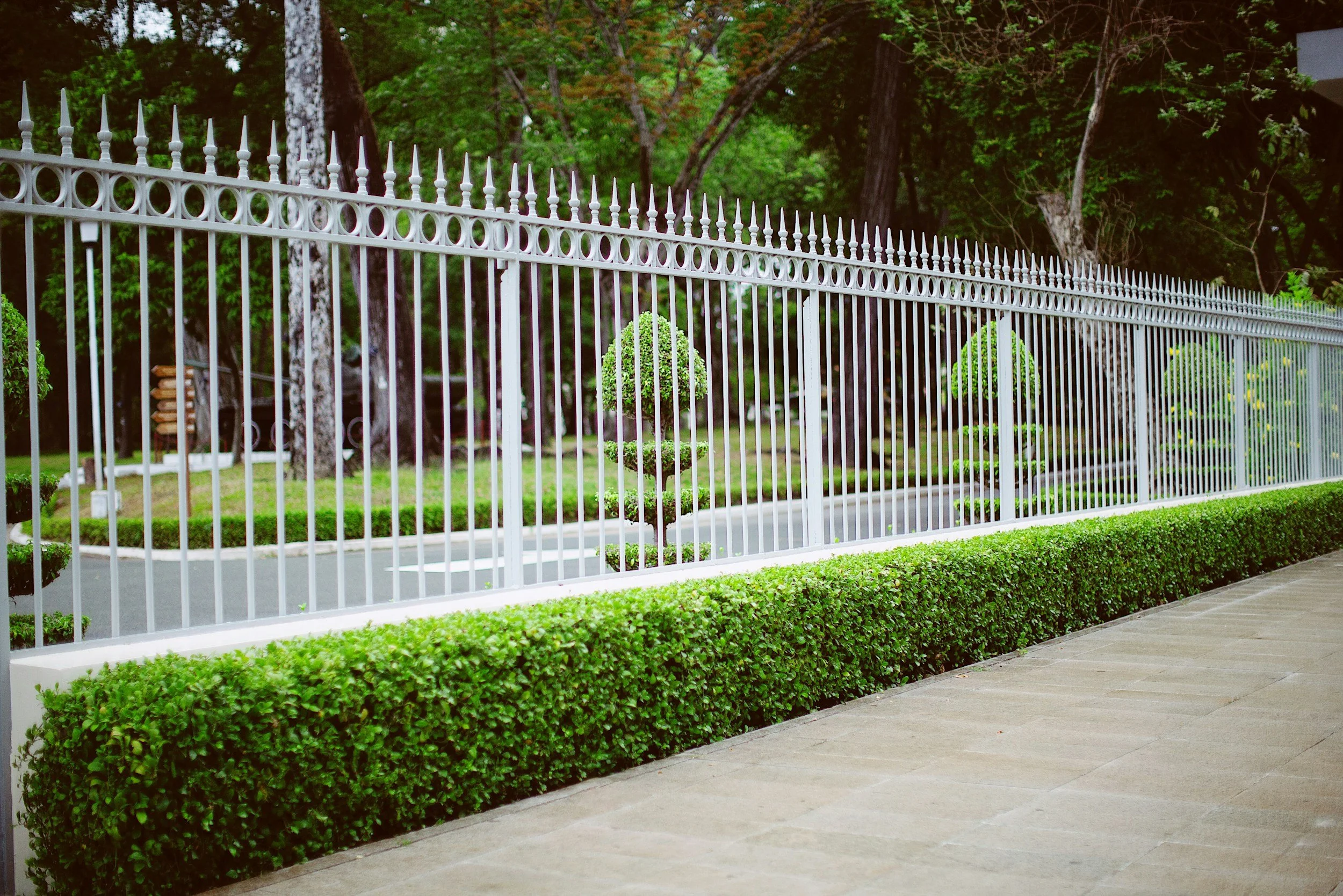 A white metal fence with pointed tips, running alongside a green hedge and sidewalk. Behind the fence, there are manicured bushes and tall trees with lush green foliage.