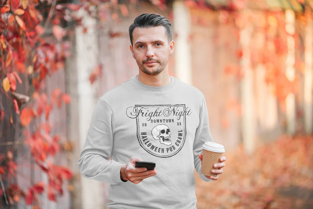 Man in a light sweater with a Halloween design holds coffee and phone, standing by a fence adorned with autumn leaves. The atmosphere is casual and cozy.
