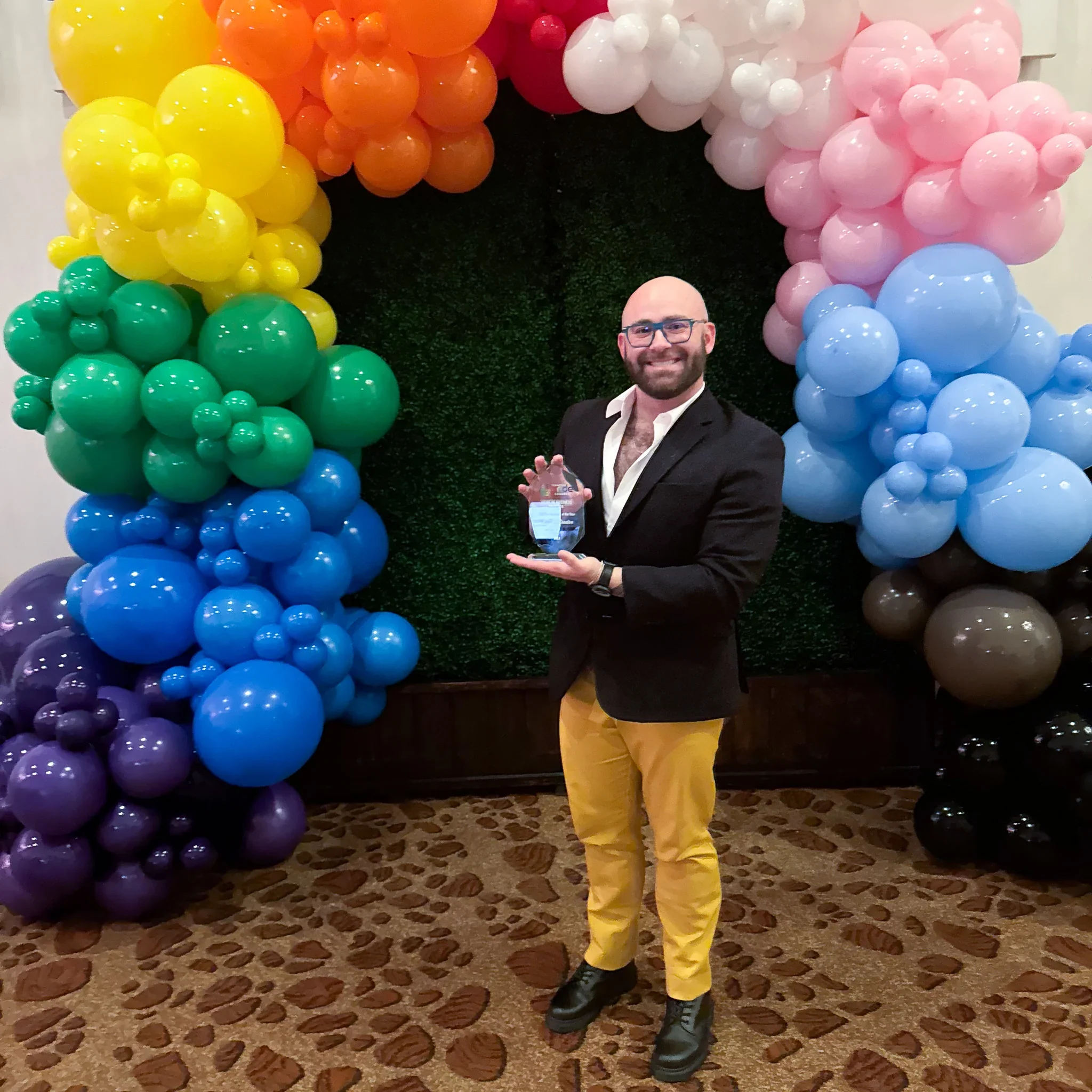 Co-founder of Music City Creative, Steven Romeo, with glasses and a beard smiles while holding a glass award. They stand in front of a vibrant rainbow balloon arch, exuding a celebratory and joyful mood.