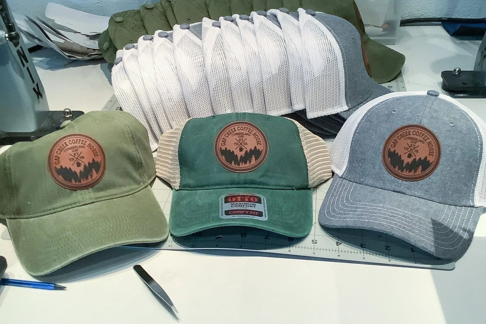 Three trucker hats with leather patches featuring the logo for Gap Creek Coffehouse are displayed on a table. Behind them are more hats stacked neatly, showcasing organization.