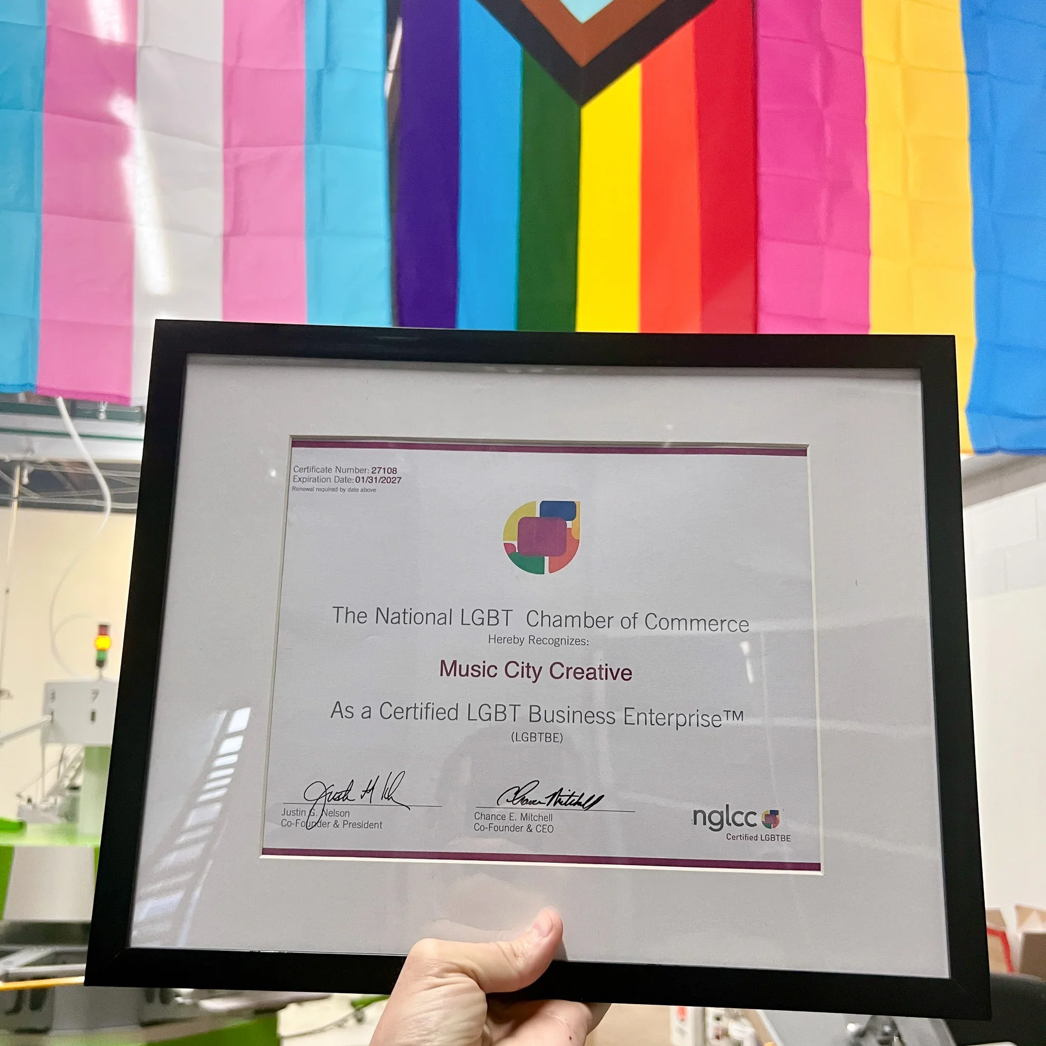 A framed certificate from the National LGBT Chamber of Commerce recognizing Music City Creative is held up. A vibrant Pride flag hangs in the background.