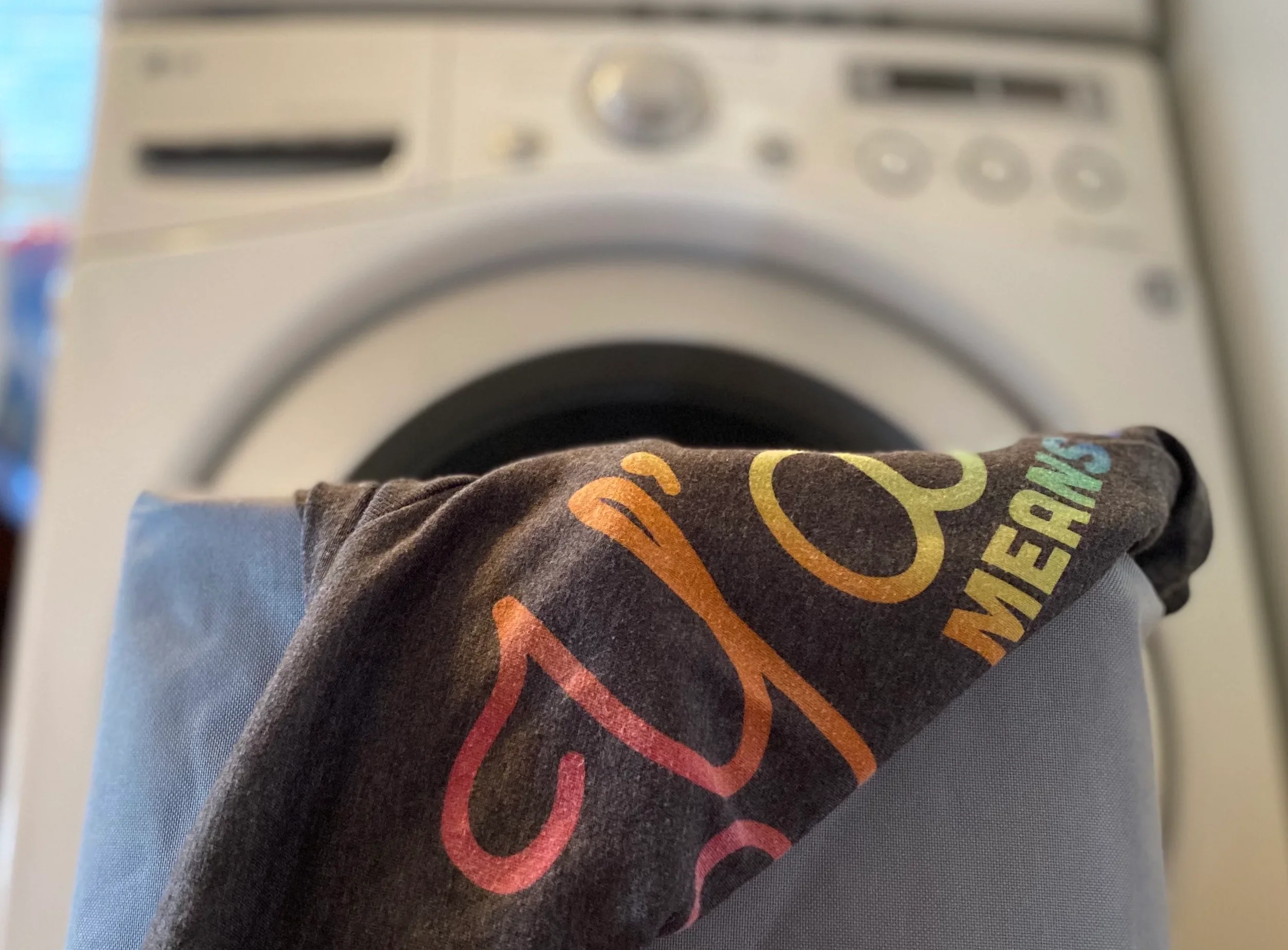 Y'all Means All t-shirt sitting in laundry basket in front of a washing machine.