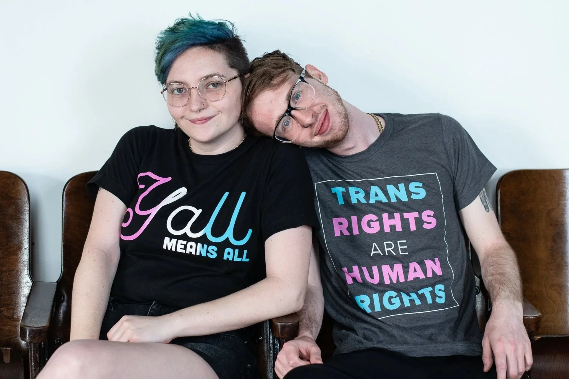 Two people sitting down wearing t-shirts promoting Transgender rights. One shirt says "Y'all Means All" in the Transgender flag colors and the other shirt says "Trans Rights Are Human Rights."