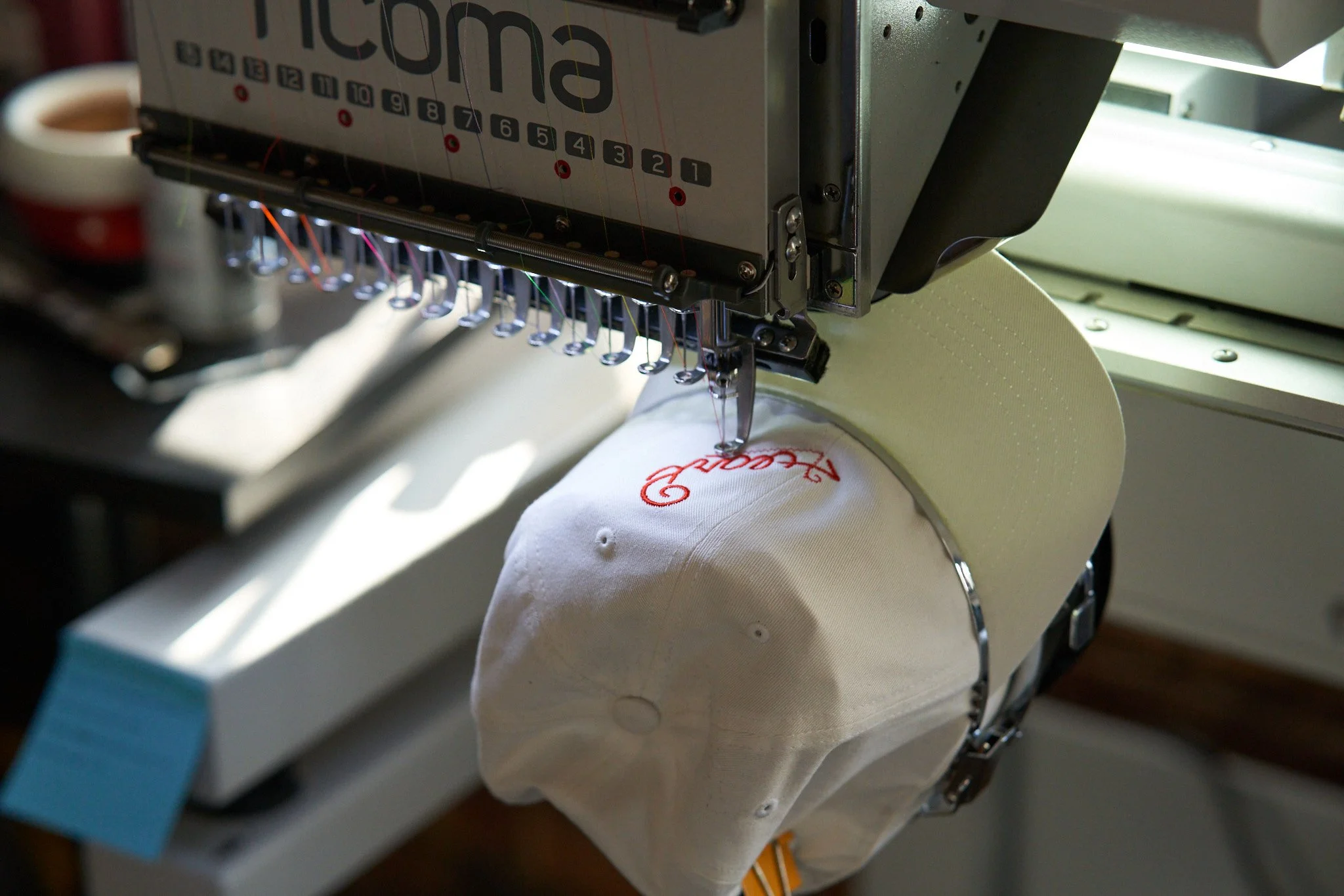 A white cap is being embroidered with red thread using an industrial embroidery machine. The brand name is partially visible, creating a focused and professional tone.