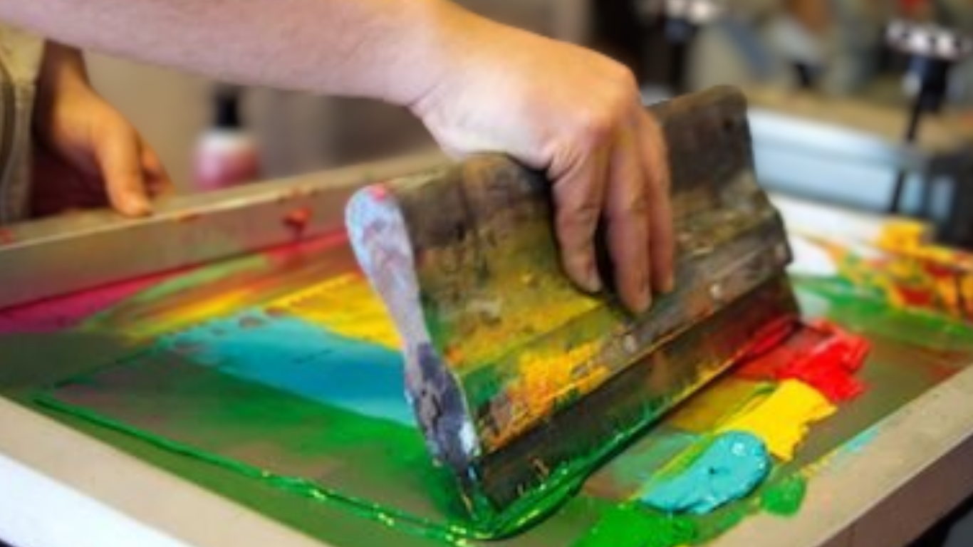 A hand using a wooden squeegee spreads vibrant inks in green, yellow, blue, and red on a screen for printing, evoking creativity and craftsmanship.