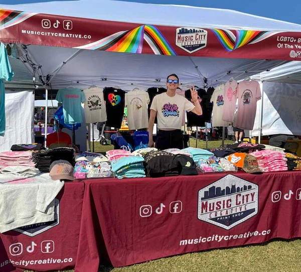 Person stands in a booth with a red "Music City Creative" banner, displaying colorful shirts and hats. The booth has rainbow accents and a friendly vibe.
