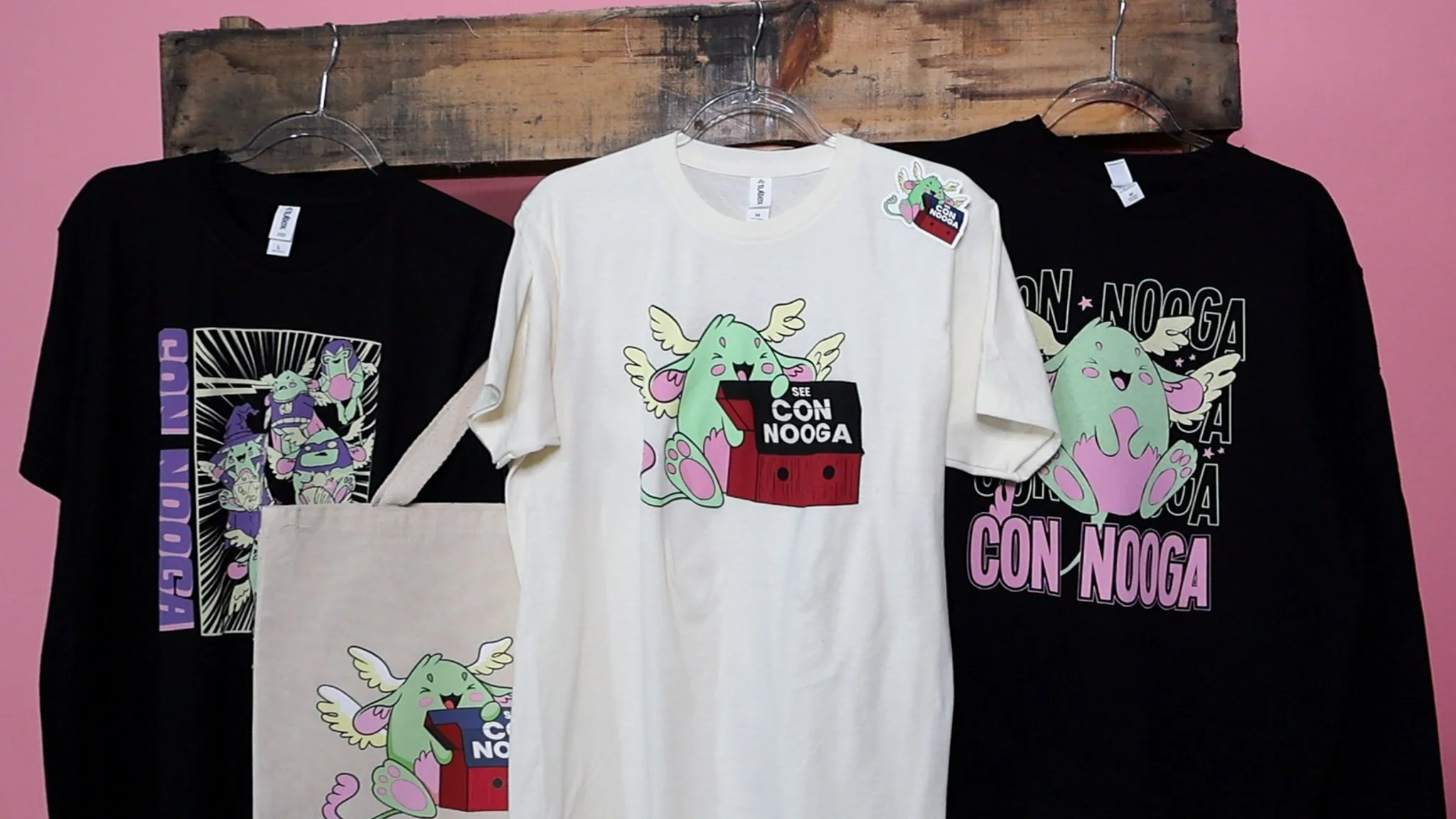 Four pieces of clothing, including two black and one white T-shirt and a tote bag, display a green, winged character with text "Con Nooga" against a pink backdrop.