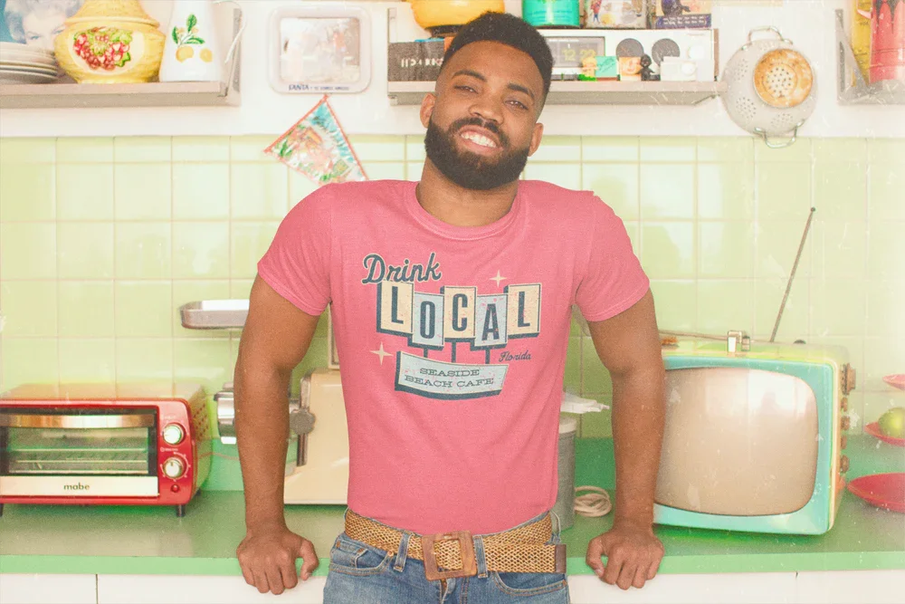 Smiling man in a pink "Drink Local" shirt stands in a retro kitchen with mint green tiles, a red toaster oven, and a vintage TV, conveying a nostalgic vibe.