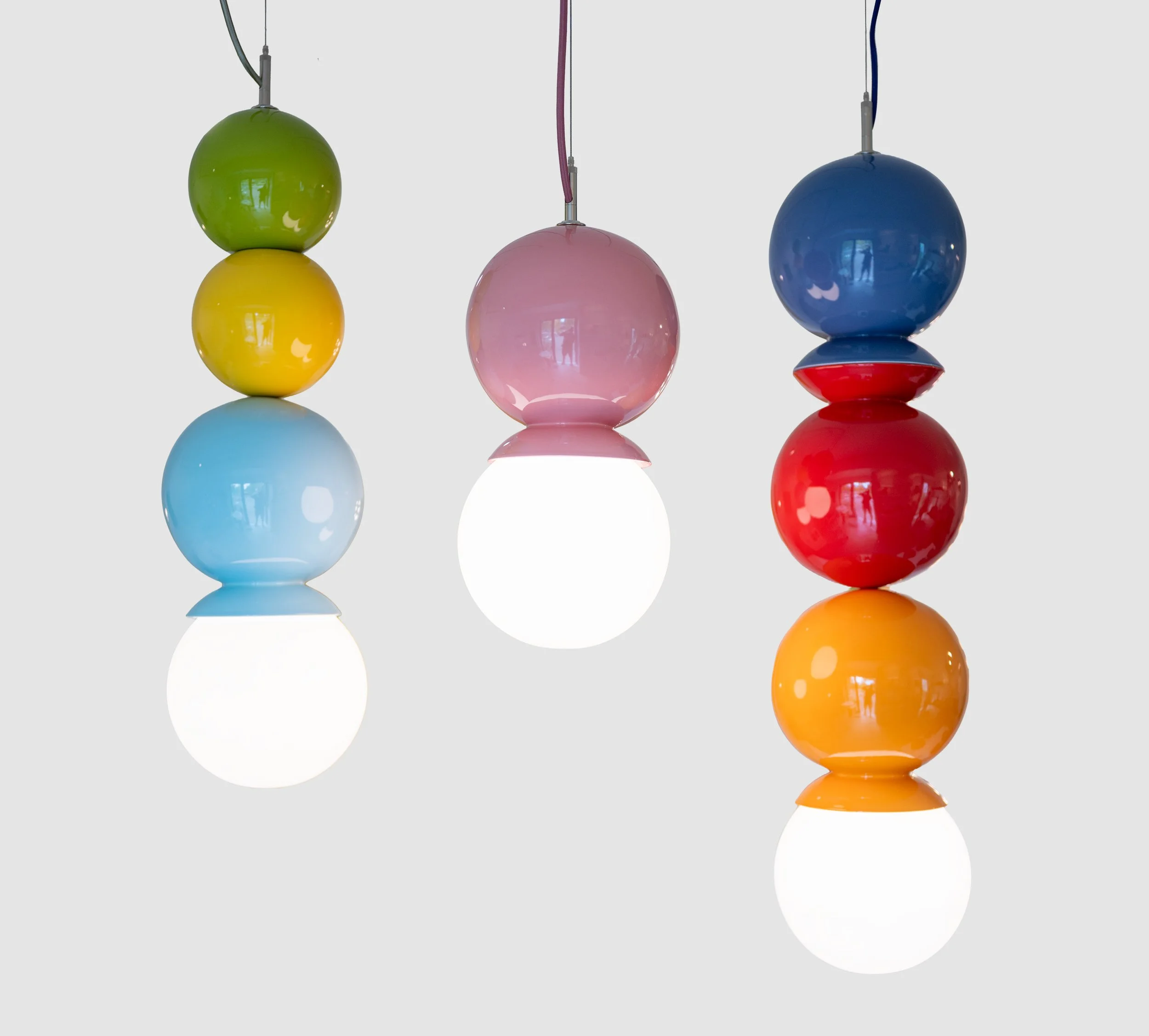 Three ceramic Dot Dot Dot pendants side by side: olive, sun yellow and light aqua; pink; and purple, sumac red and orange peel, each over a lit white globe, J Schatz Studio.
