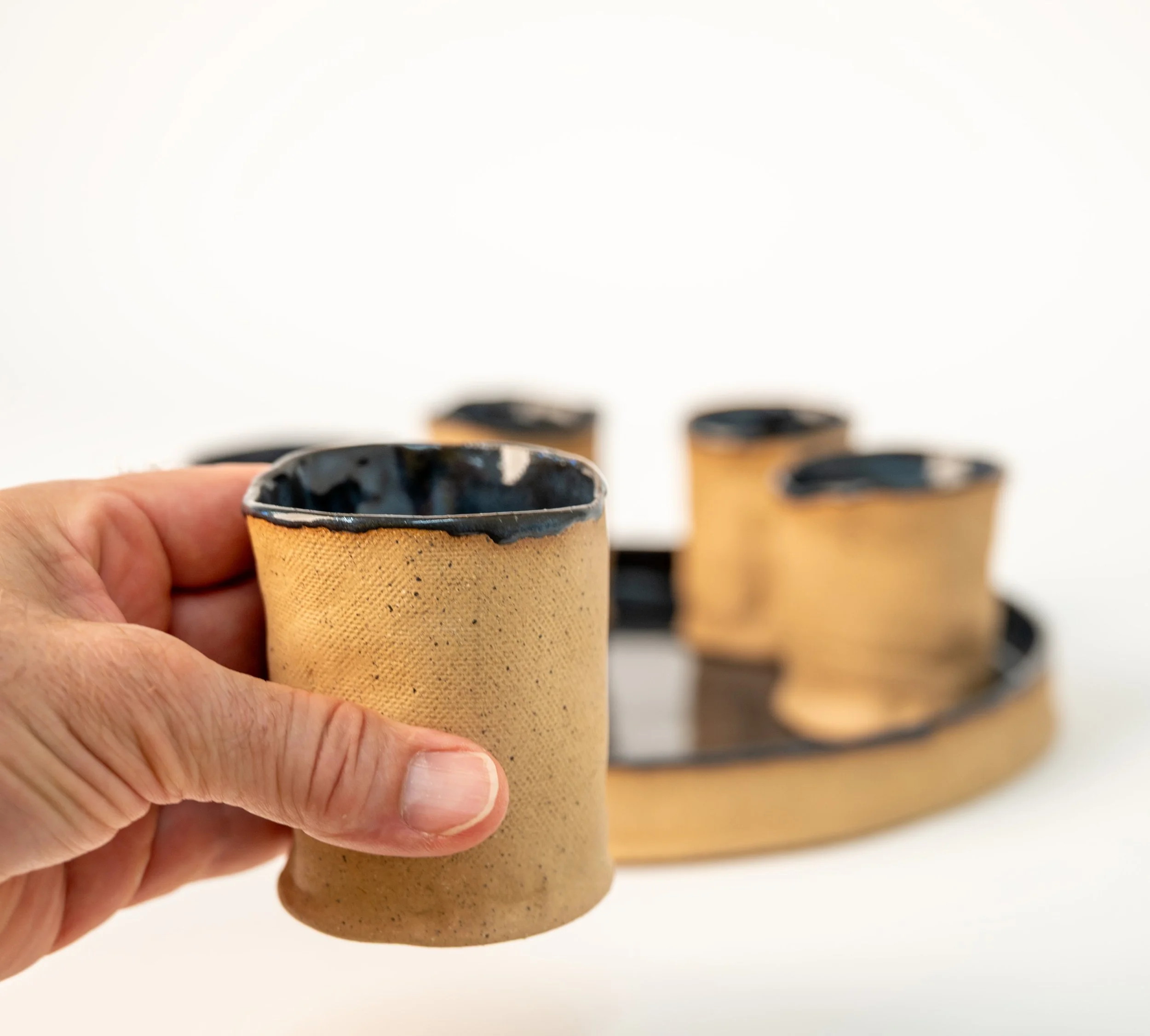 Round Tray and 6 Cups by Peter Souza Jr. — single stoneware cup held in hand showing speckled raw clay texture and glazed rim, set in background