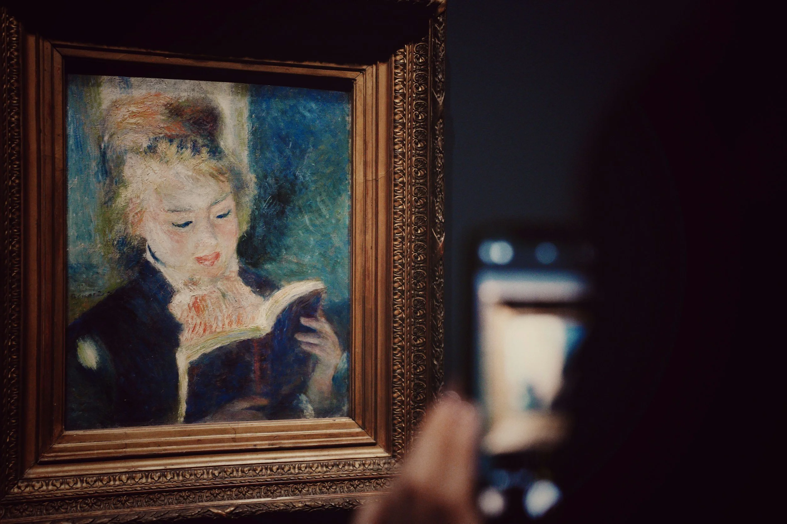 Person taking a photo of an art painting of a woman with light-colored hair reading a book, framed in an ornate gold frame.