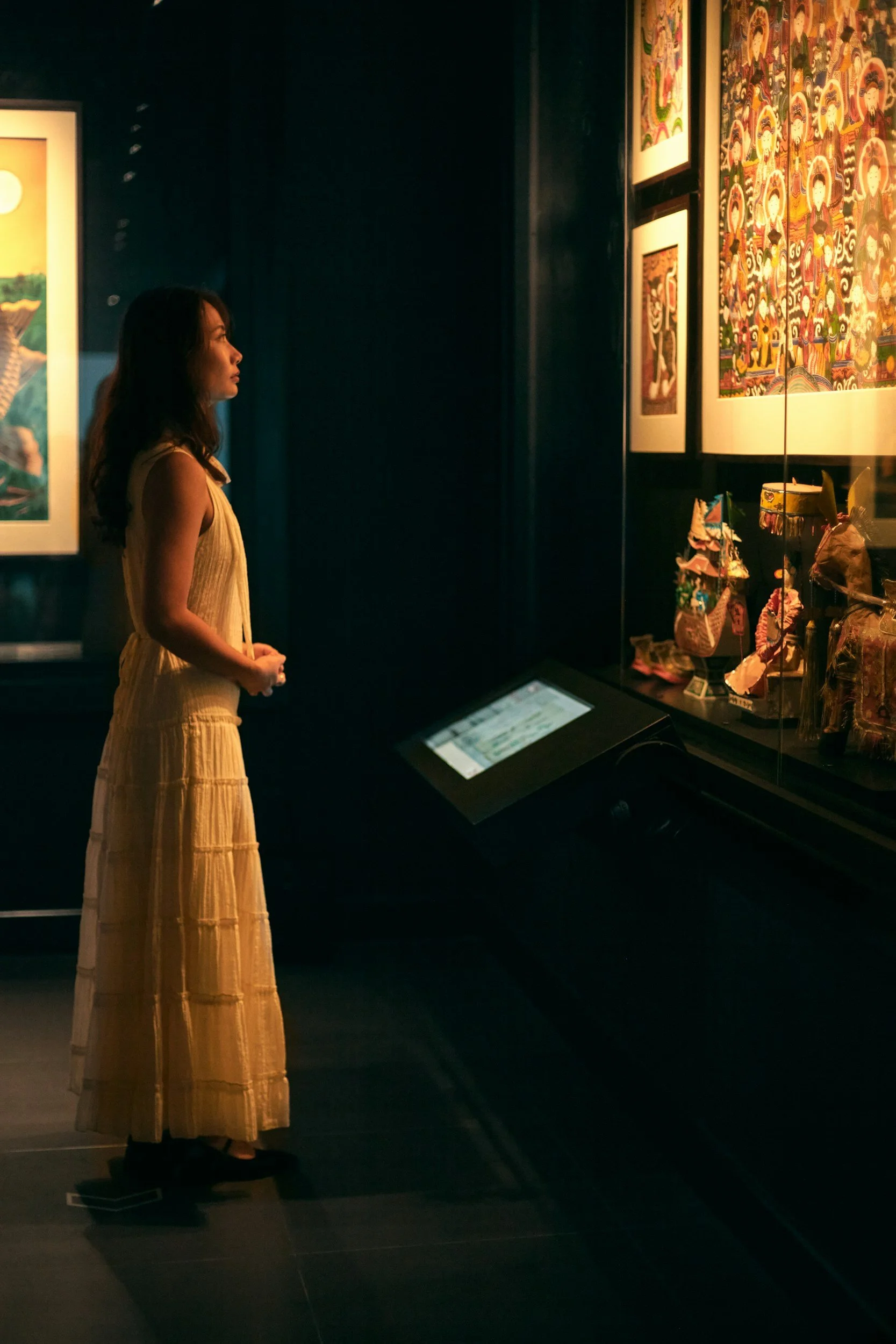 A woman in a long, sleeveless yellow dress looks at artwork displayed in a dimly lit gallery, with another larger piece of colorful art on the wall behind her.