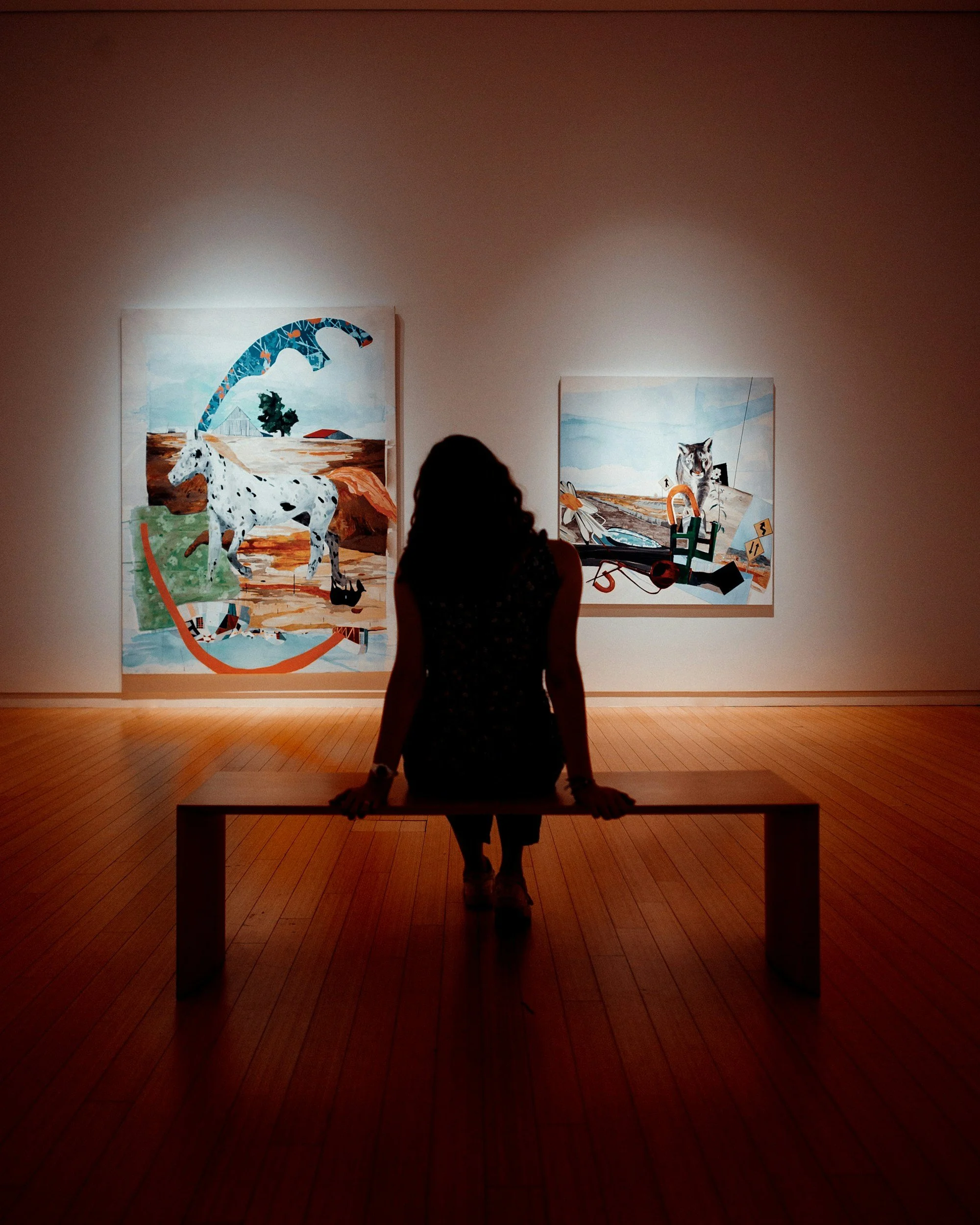 A woman sitting on a bench in an art gallery, viewing two colorful abstract paintings featuring animals on a white wall.
