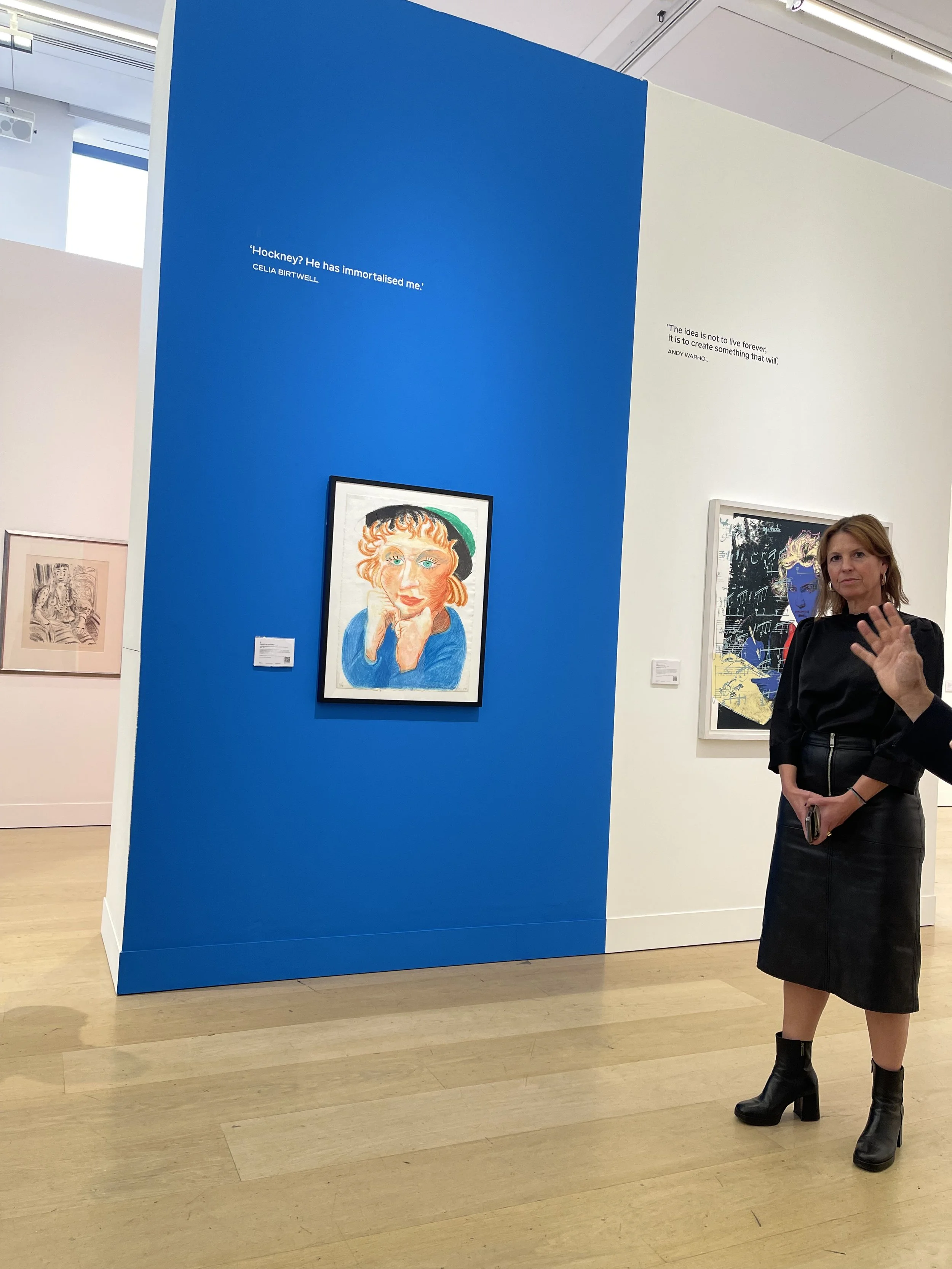 A woman with blonde hair wearing a black dress and black boots standing in an art gallery, gesturing with her hand. Behind her are framed artworks, with a blue wall displaying quotes about art.