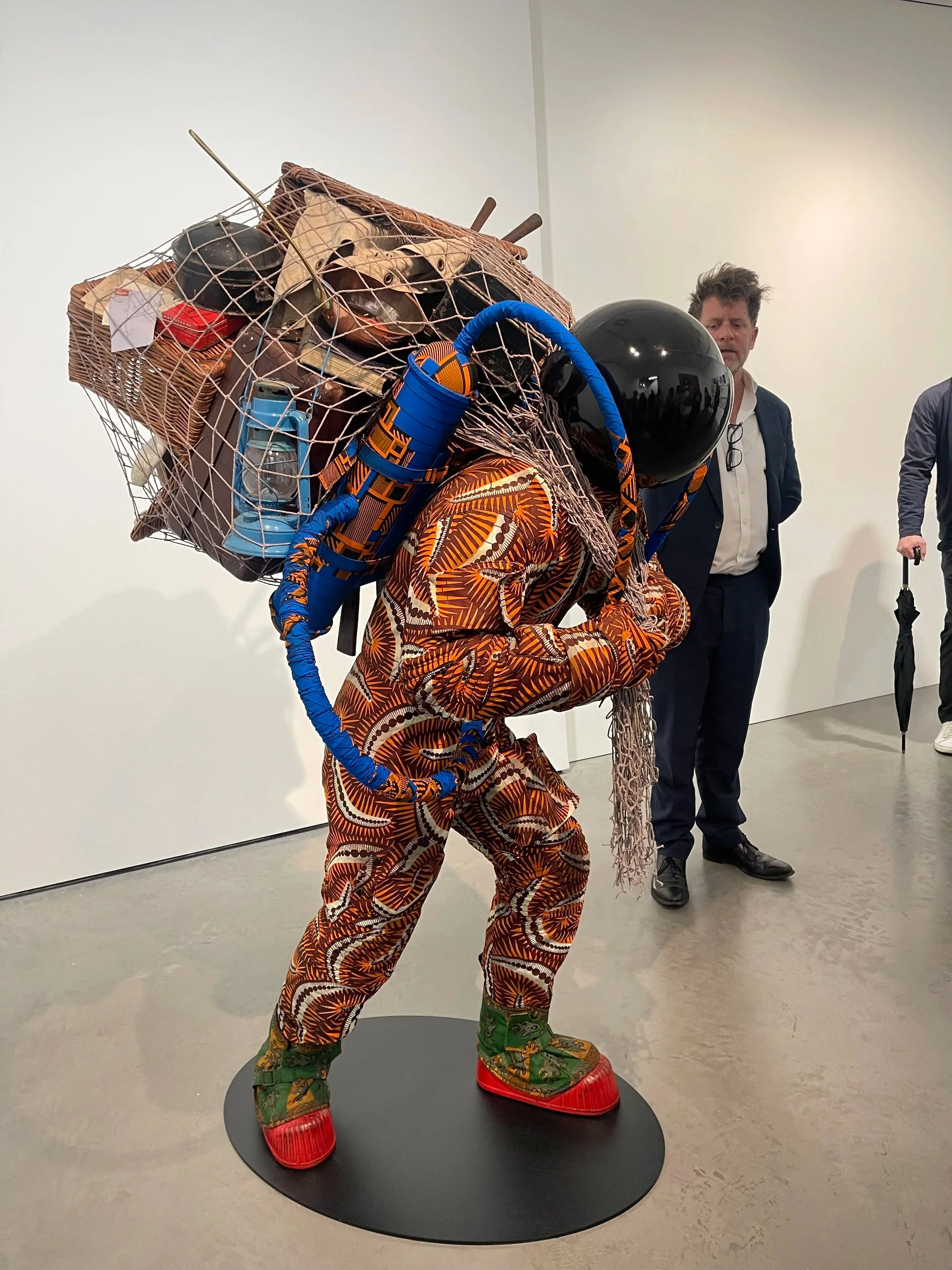 Artist wearing a brightly patterned jumpsuit with a large, complex headpiece made of various objects, including a black helmet, metal netting, a basket, and other miscellaneous items, on display at an art gallery.