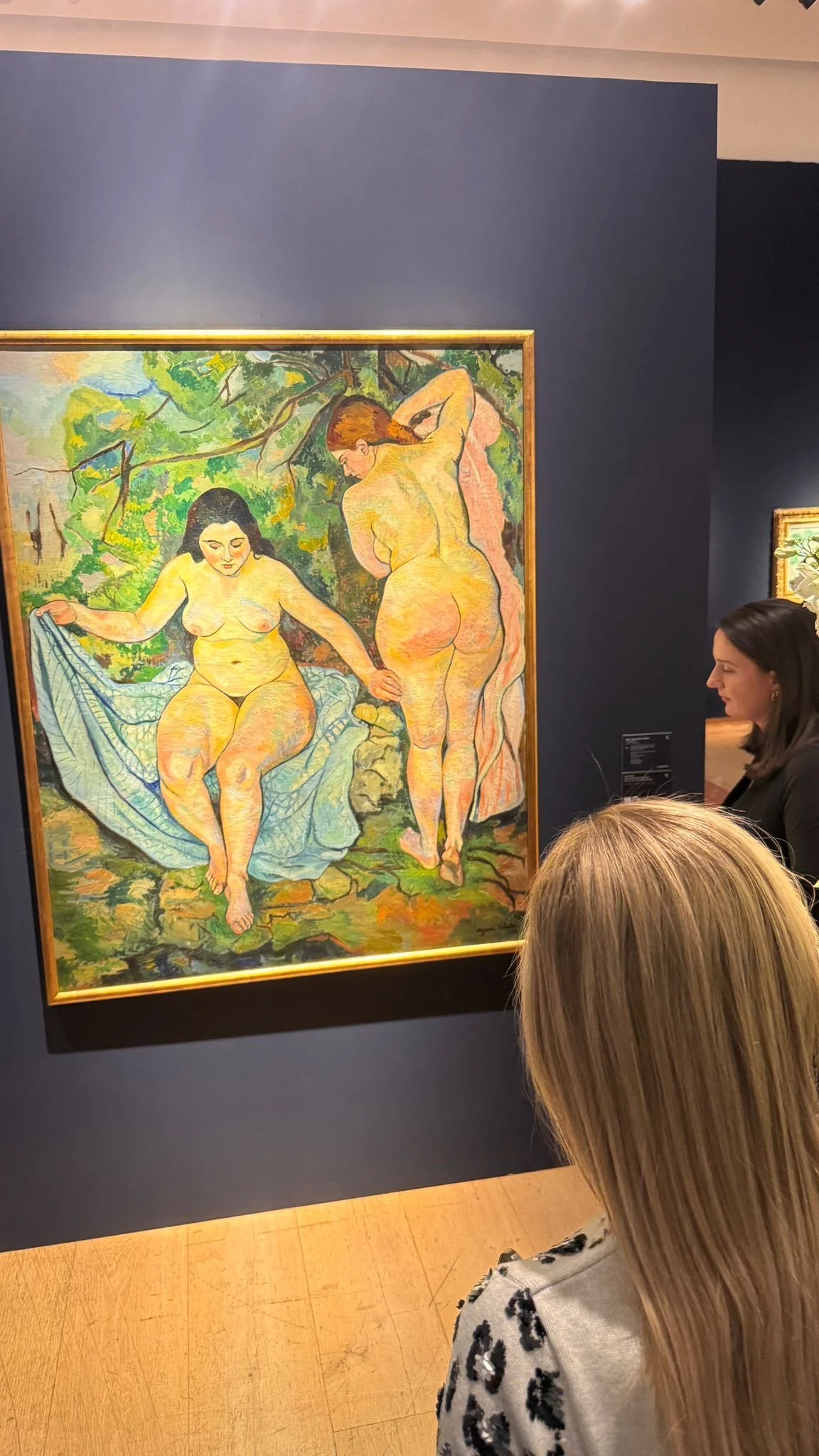 People observing a painting of two nude women in a lush outdoor setting, with one seated and the other standing, holding a downy object, surrounded by vibrant greenery.