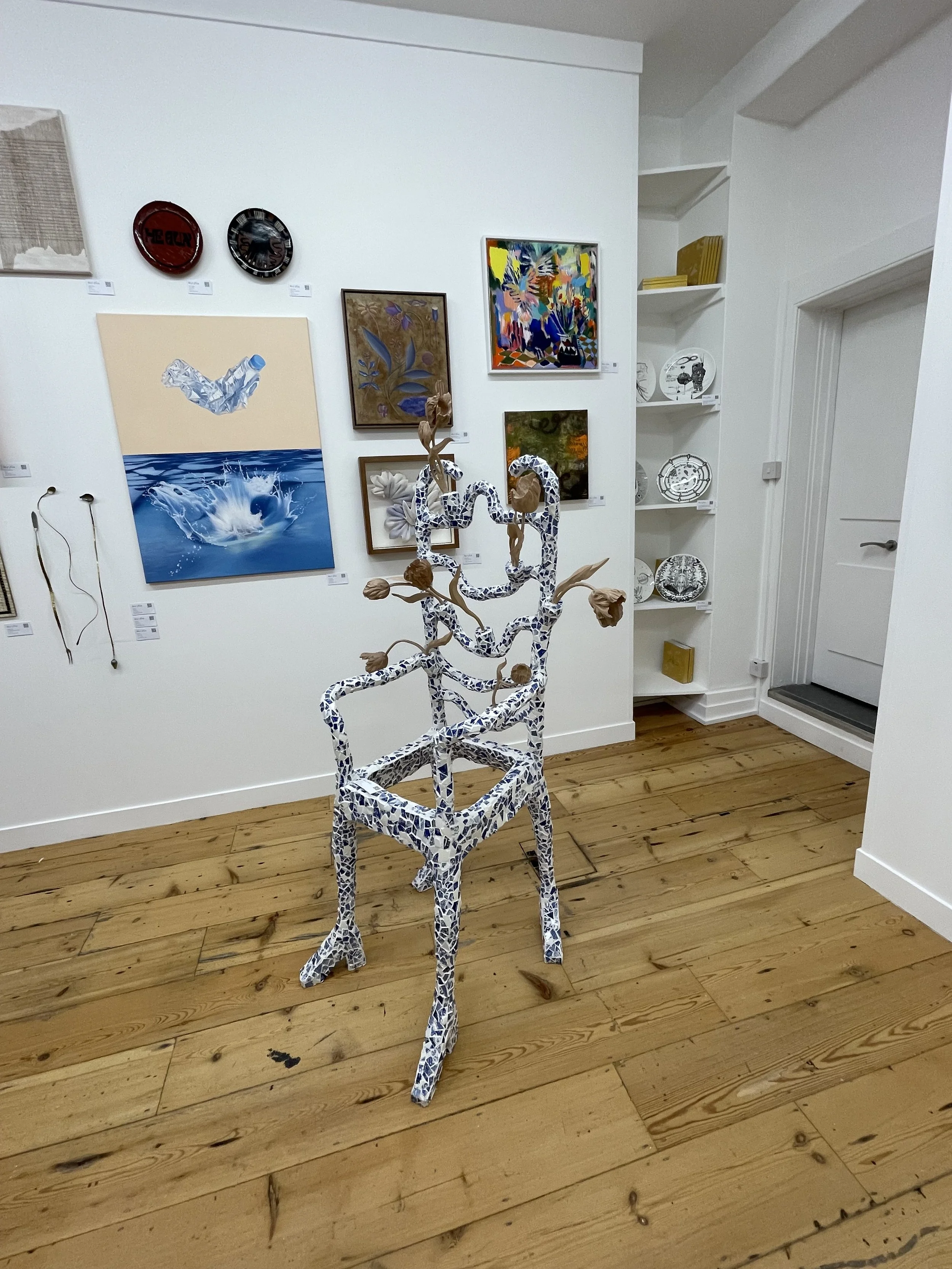 Art gallery with paintings and ceramic plate displays on white walls and a wooden floor, featuring a mosaic chair sculpture with a tree-like structure.