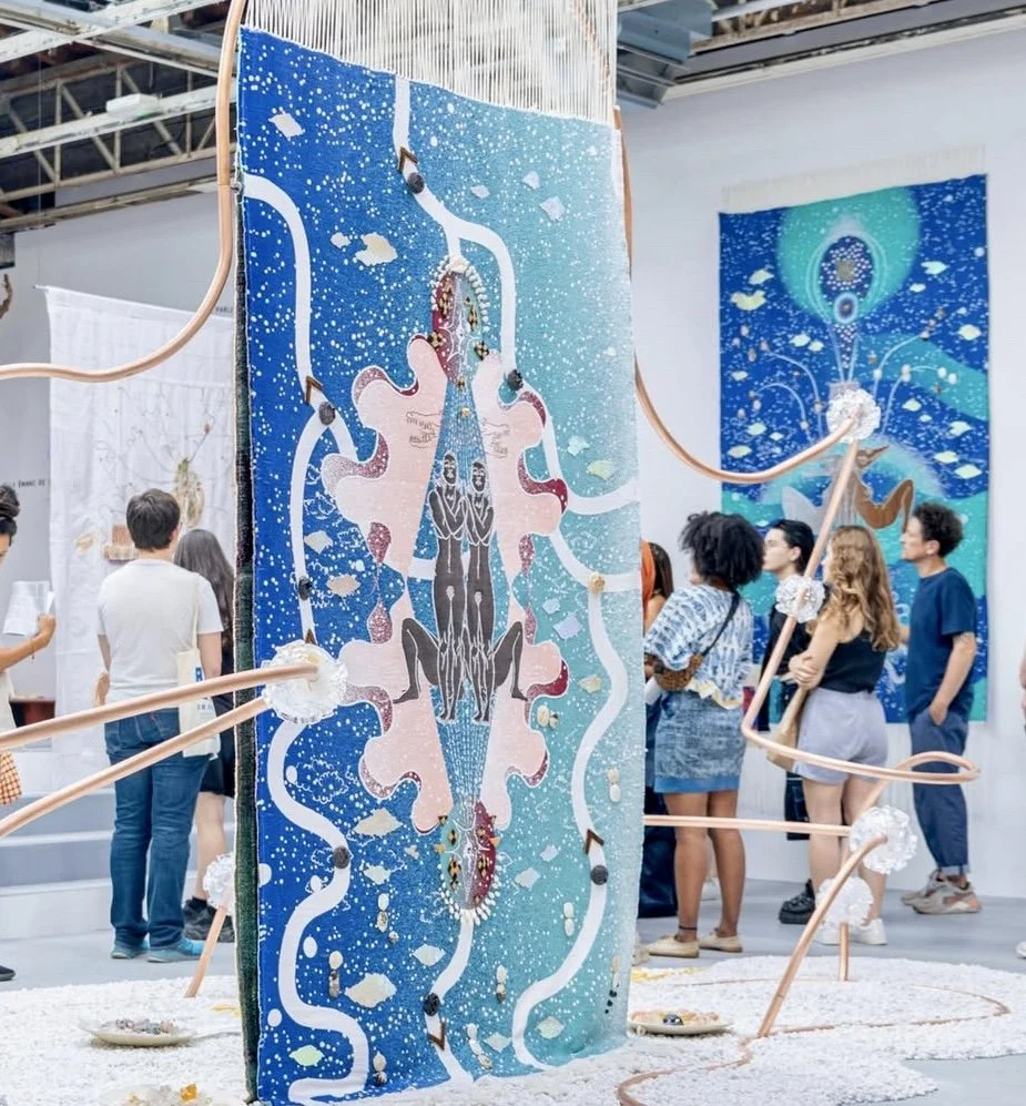 People viewing abstract artwork in an art gallery. The artwork is a large vertical textile piece with a symmetrical, colorful design featuring blue, pink, white, and black patterns, some resembling a butterfly or angel, with flowing lines and splatters. Copper tubing with white pom-poms loops around the art piece.
