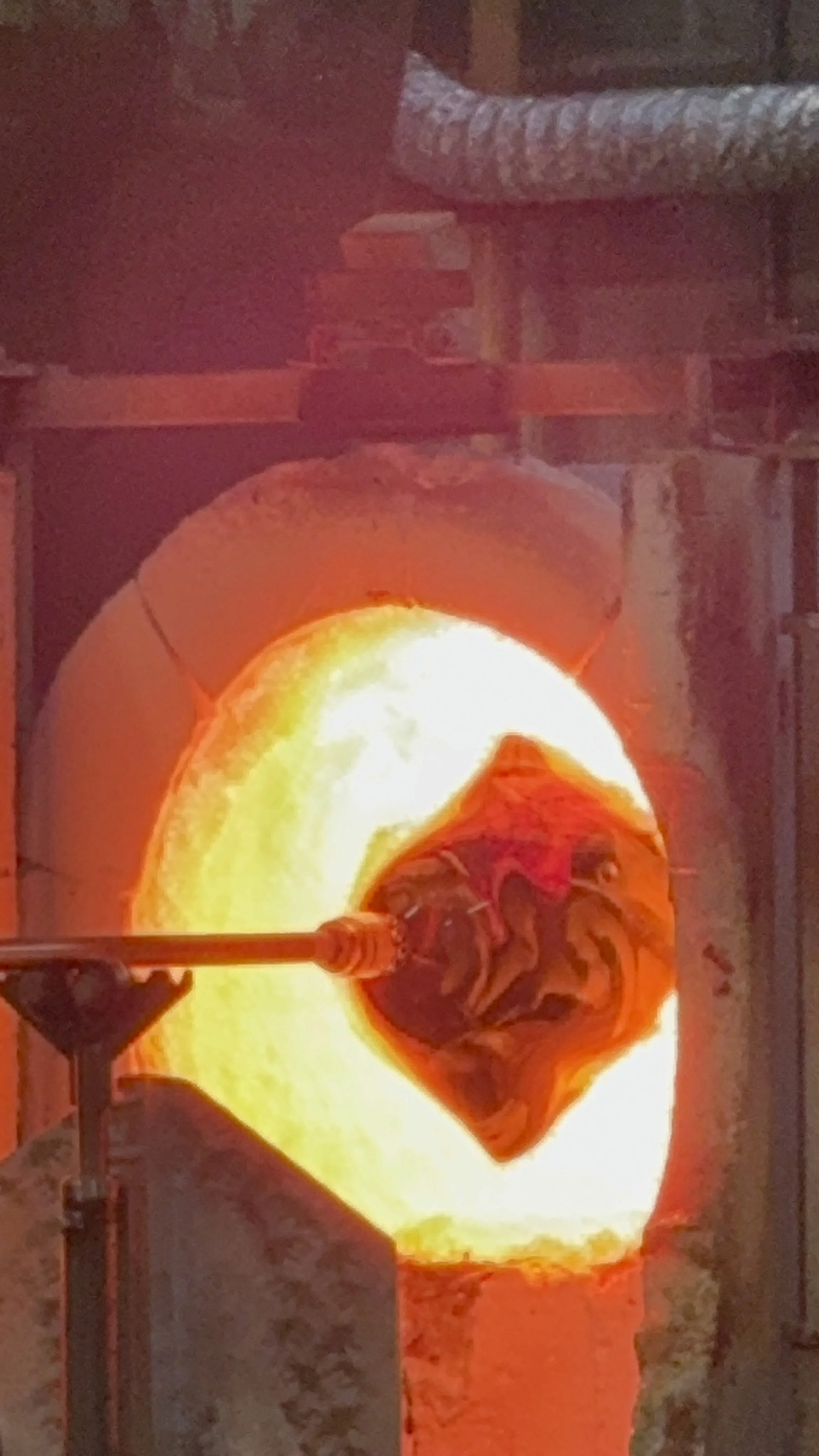 A glassblowing process with molten glass on a pipe, glowing bright orange and yellow, inside a furnace.
