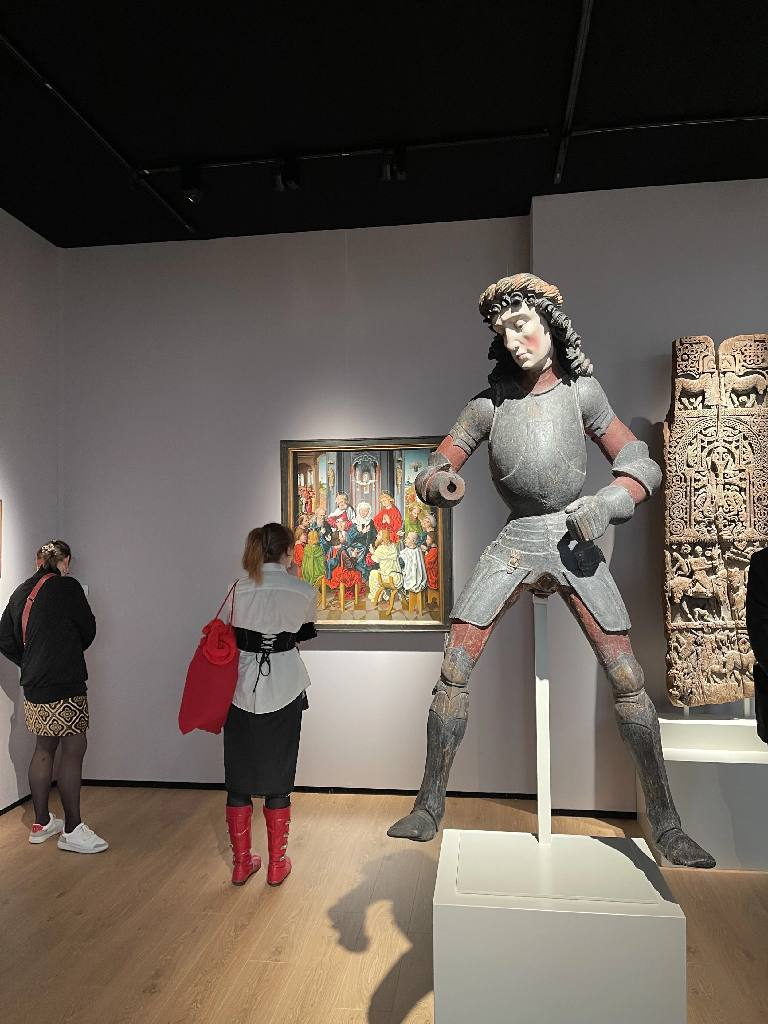 People viewing artwork, a large medieval-style armored figure statue in a museum, with a colorful painting and carved wooden panel behind.