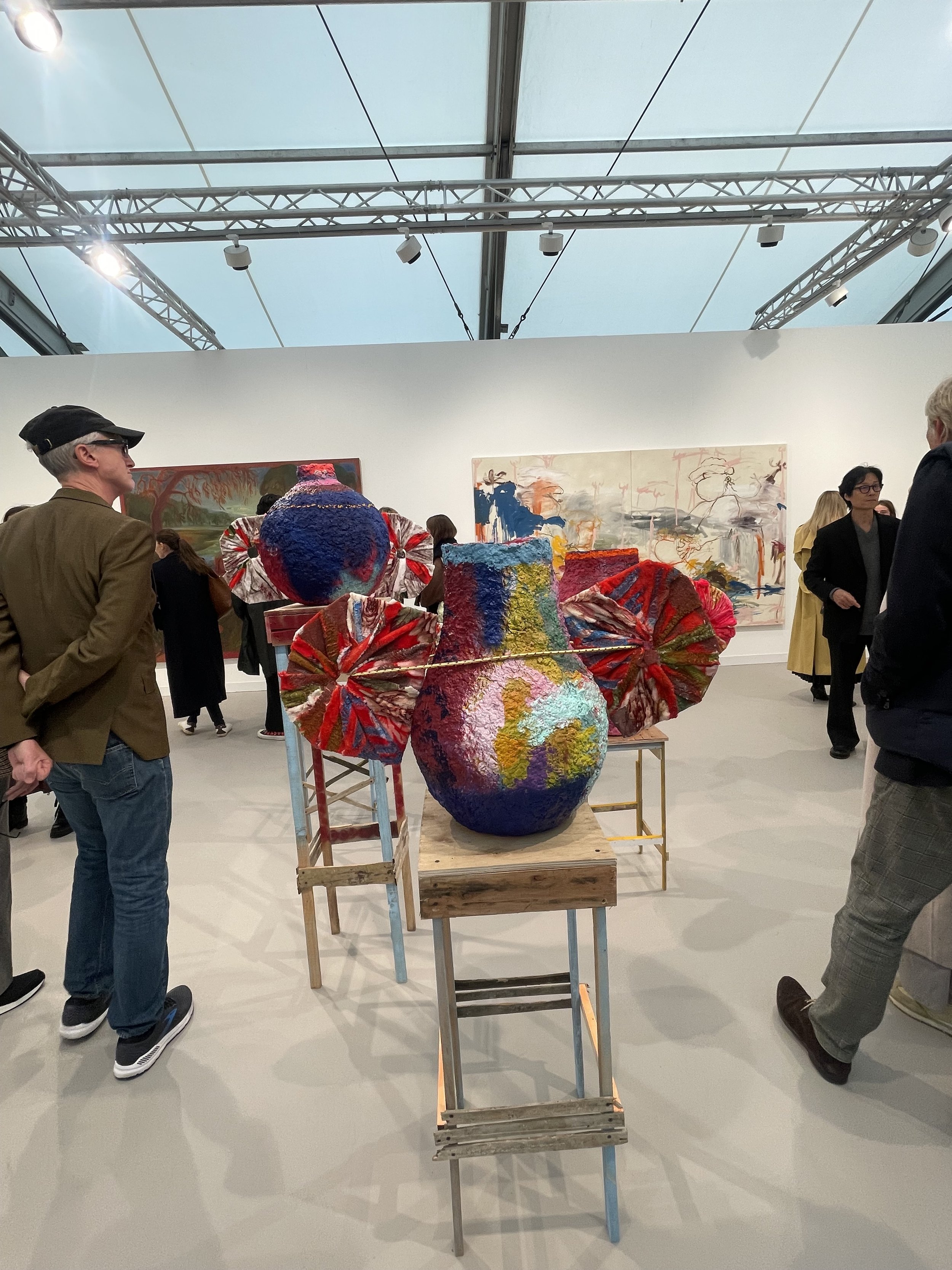 Colorful ceramic vases with textured and ribbon-like elements displayed on a wooden table at an art gallery, with viewers looking at the artwork and abstract paintings on the wall in the background.