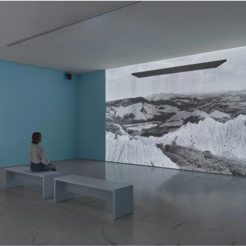 A woman sitting on a white bench in an art gallery, facing a large black and white landscape projection on the wall, with a blue wall and ceiling.