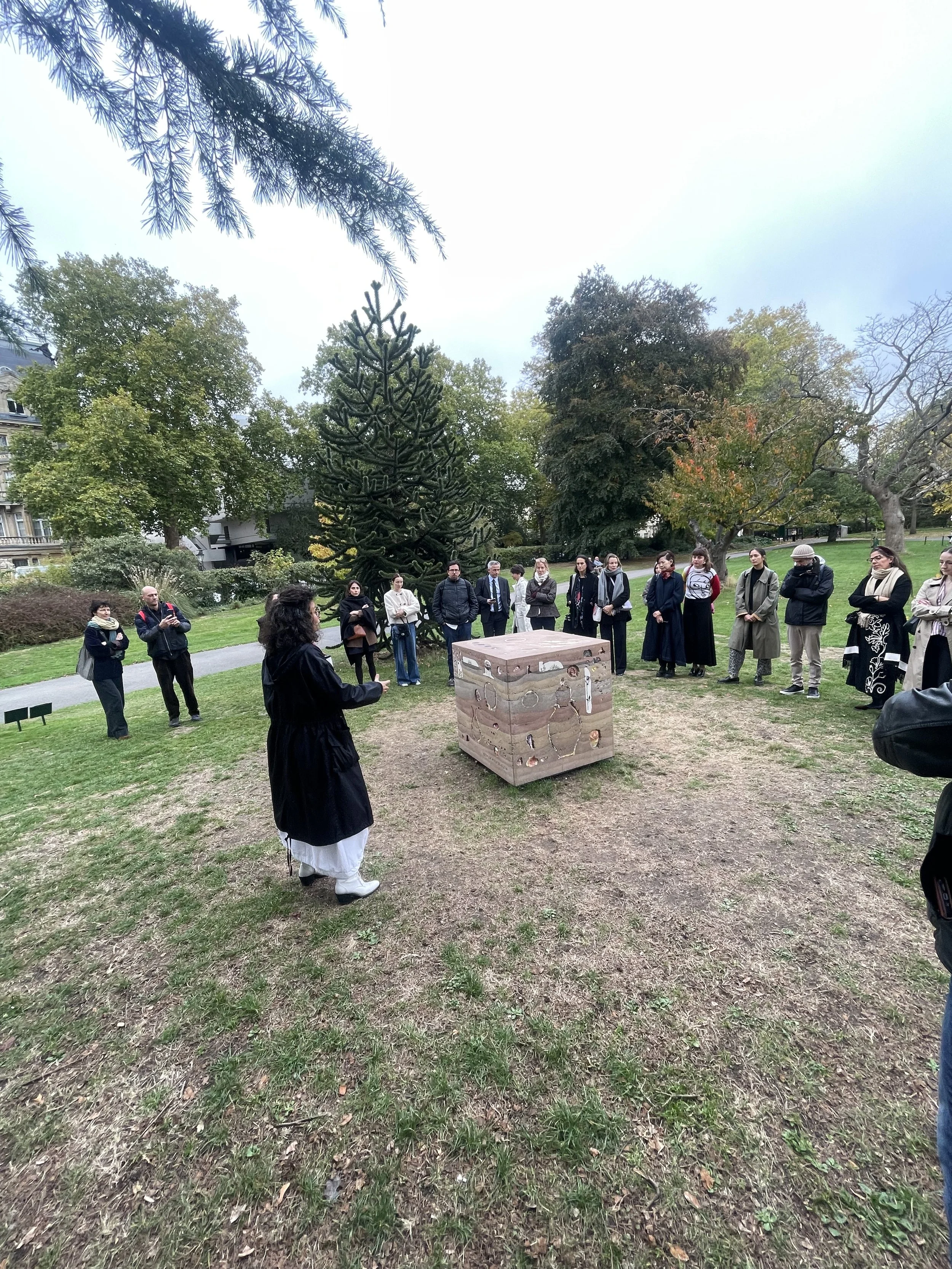 A group of people gathering outdoors in a park-like setting, listening to a woman speaking near a decorated wooden box, with trees and a grassy area in the background.