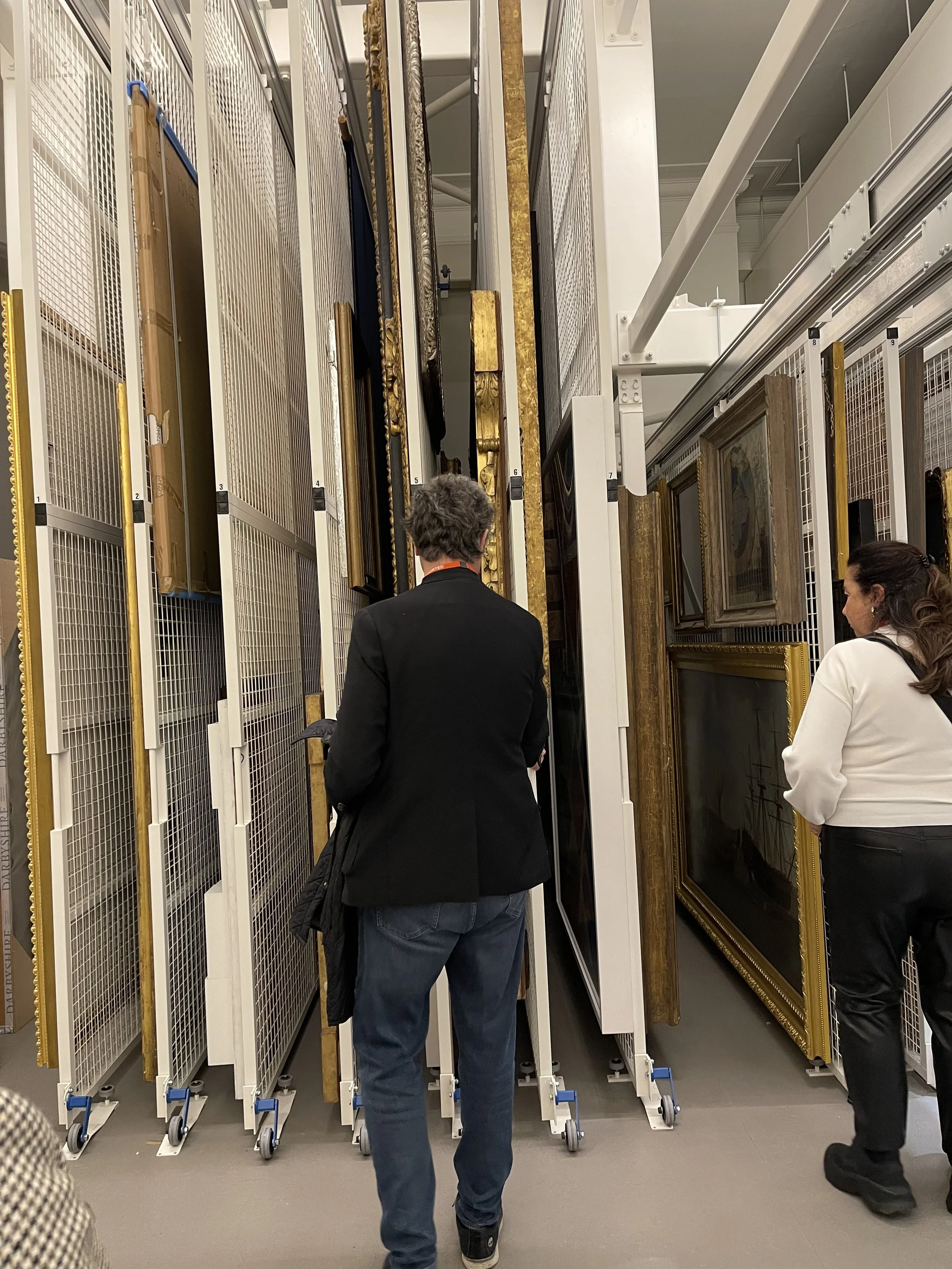 Two people viewing a tall, narrow display of framed artwork or mirrors in an art storage or gallery setting.