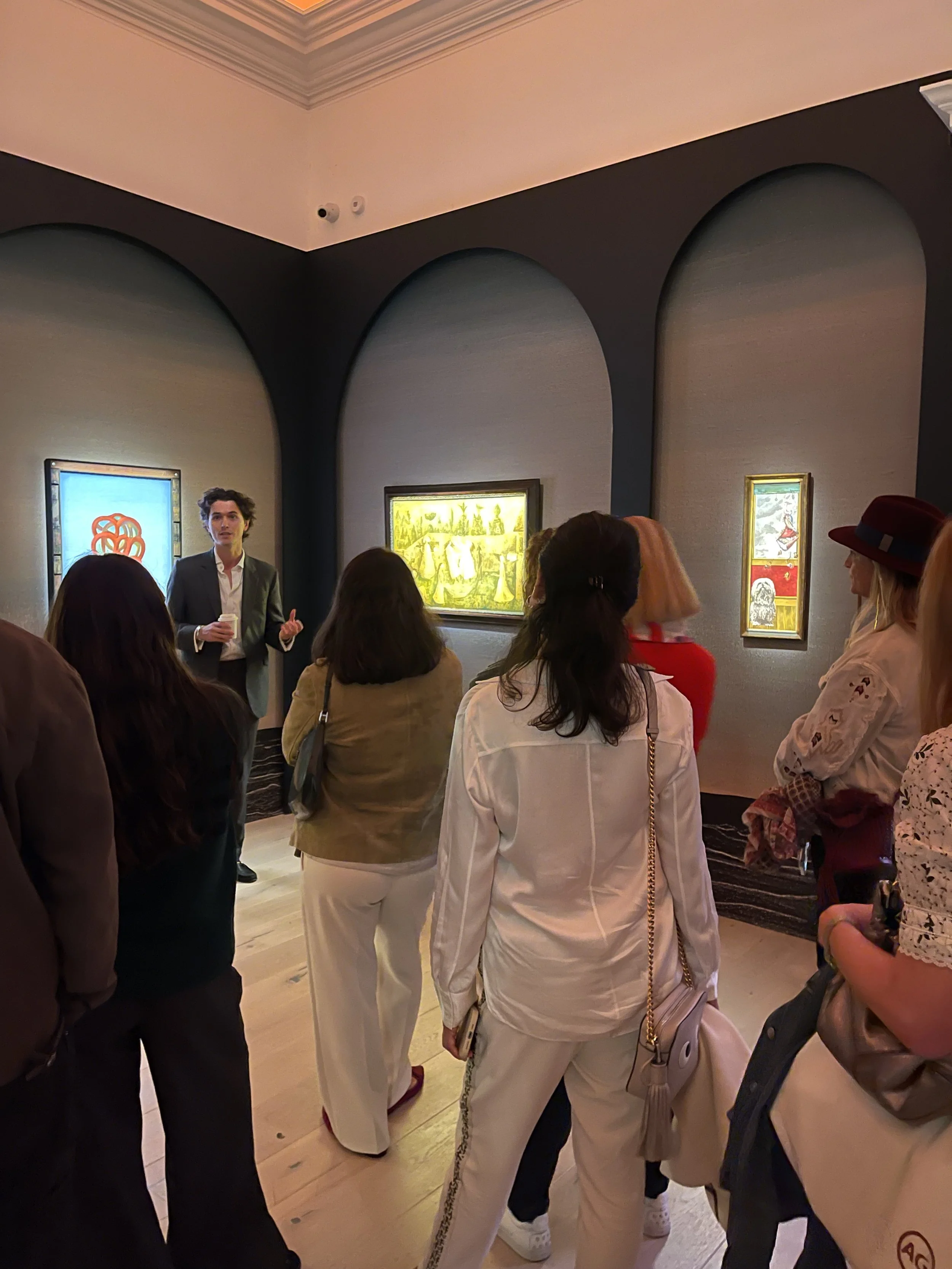 People attending an art tour or lecture in an art gallery, listening to a man in a suit explaining ART in front of three colorful paintings on a gray wall.