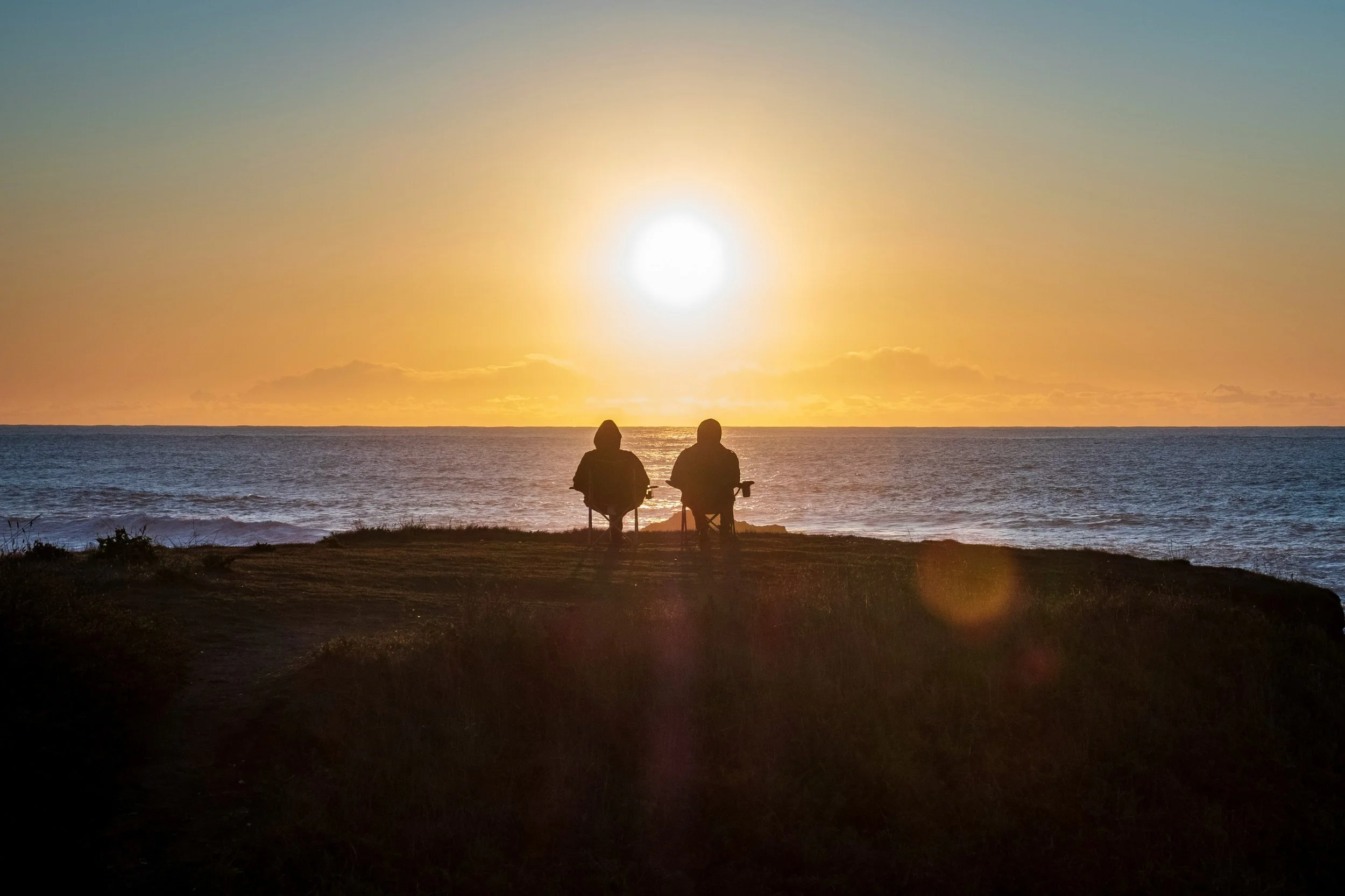 Two people sit on chairs by the ocean at sunset, facing the horizon with the sun setting over the water.