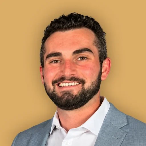 Portrait of a man with dark hair, beard, wearing a light blue suit jacket and white shirt, smiling against a beige background.