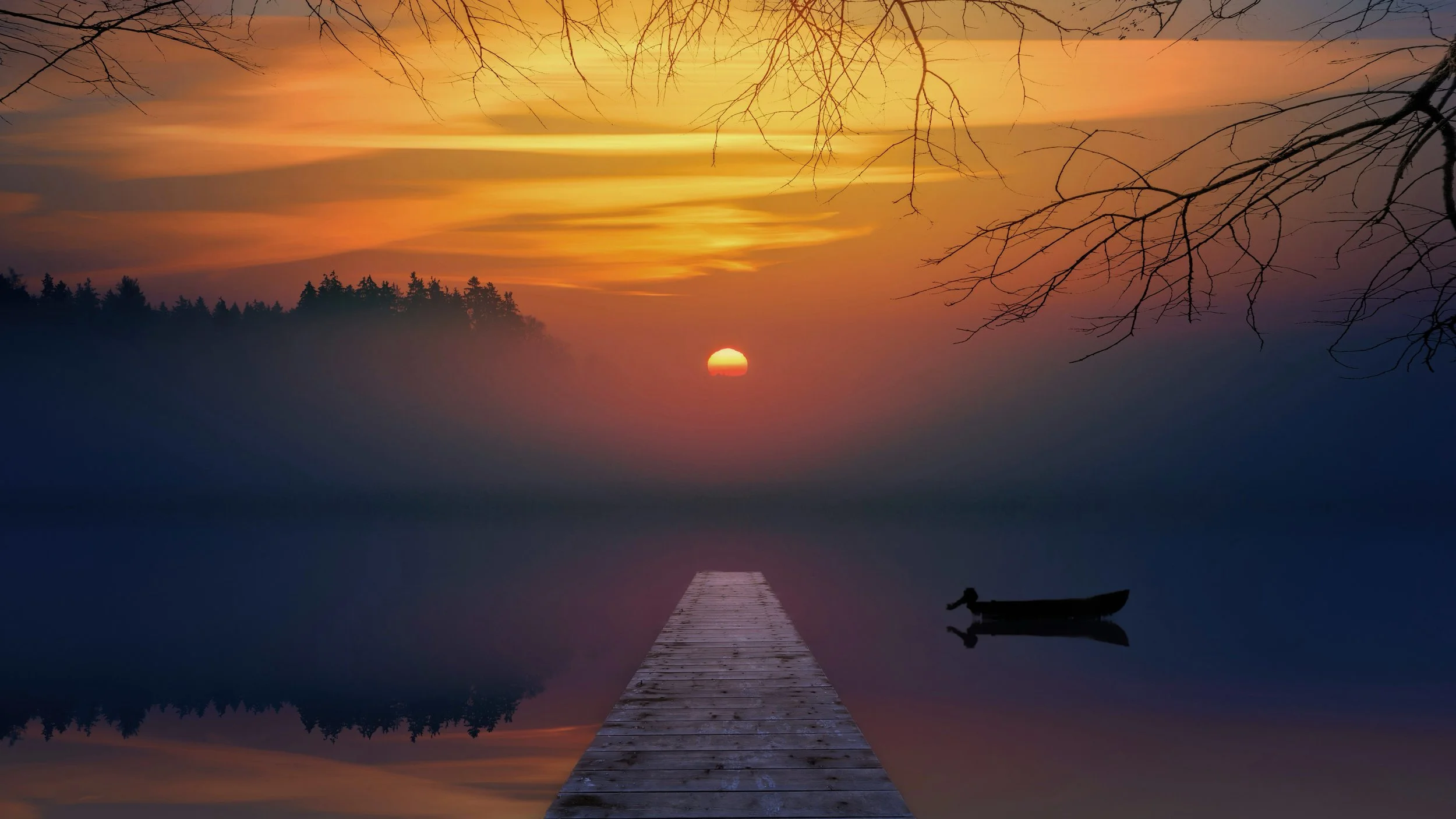 A wooden dock extends into a calm lake at sunset, with bare tree branches overhead and a silhouette of a boat on the water. The sky is filled with orange, yellow, and purple hues, and the sun is partially visible on the horizon.