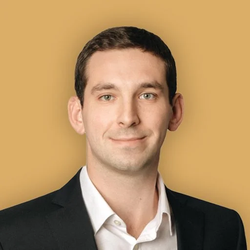 Professional headshot of a man in a suit and tie, smiling, against a beige background.
