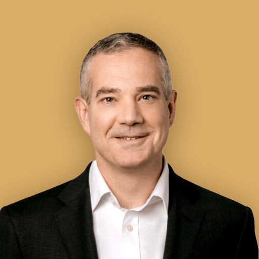 A professional headshot of a smiling man in a black suit and white shirt against a beige background.