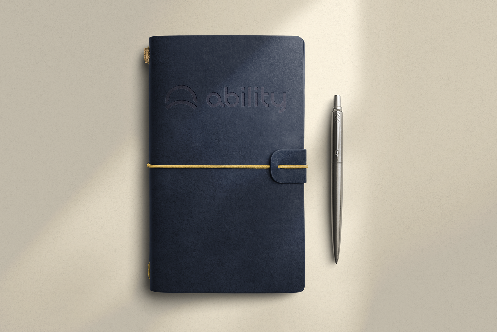 A navy blue notebook with the word 'ability' embossed on the cover, a yellow elastic band, and a silver pen placed beside it on a beige surface.