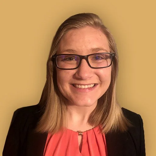 A young woman with shoulder-length blonde hair, glasses, and a bright smile, wearing a black blazer over a coral blouse, standing against a yellow wall.